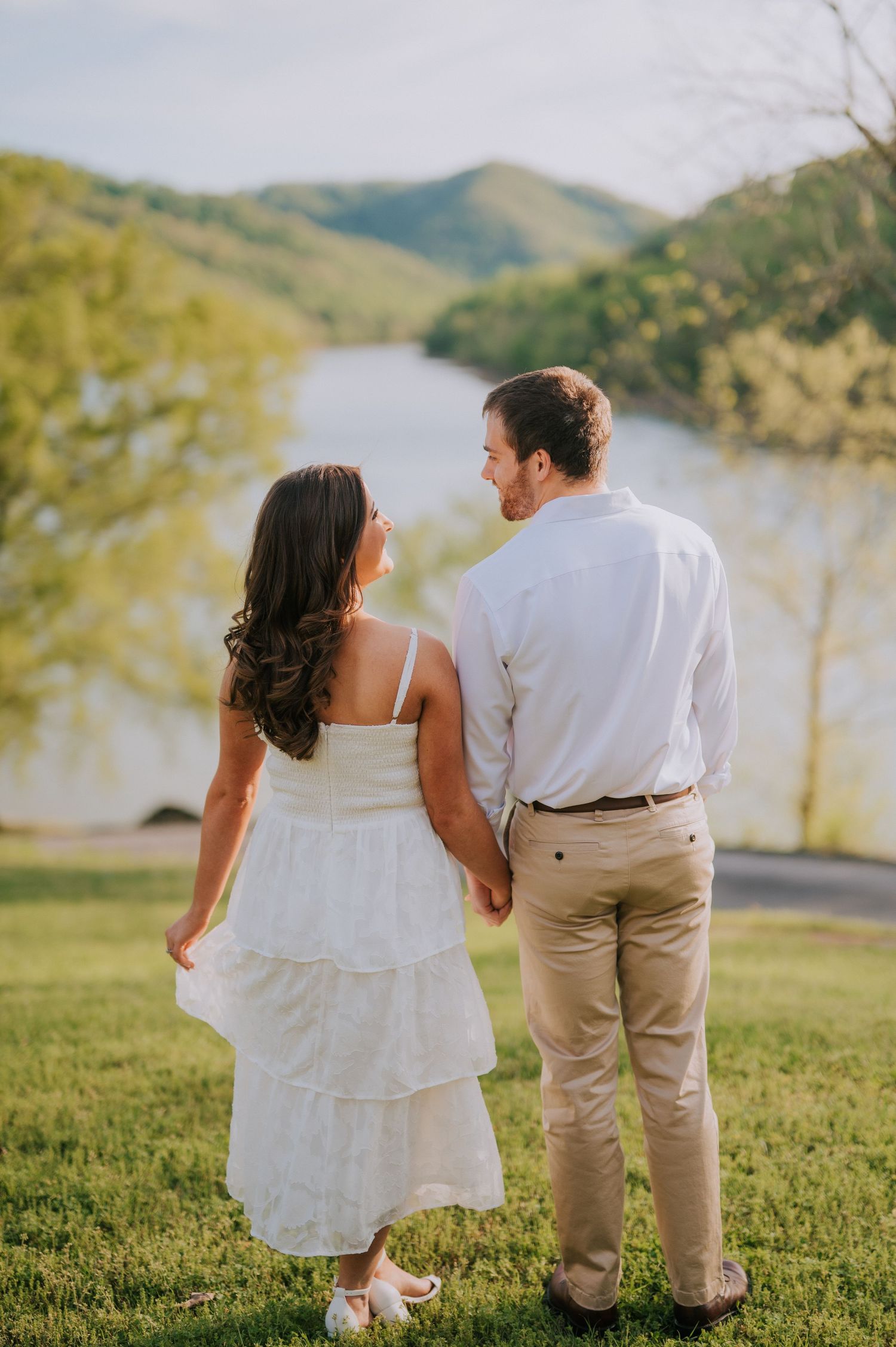 A couple in white and khaki stands together on grass overlooking a serene lake view.