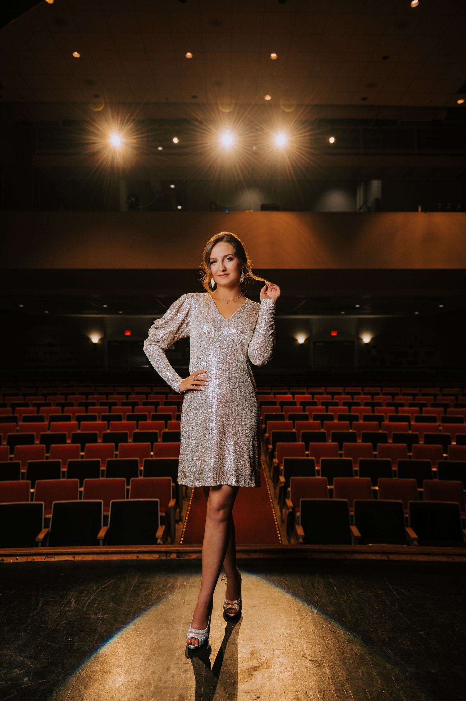 A performer in a sparkly silver dress poses confidently on an empty theater stage under dramatic lighting.