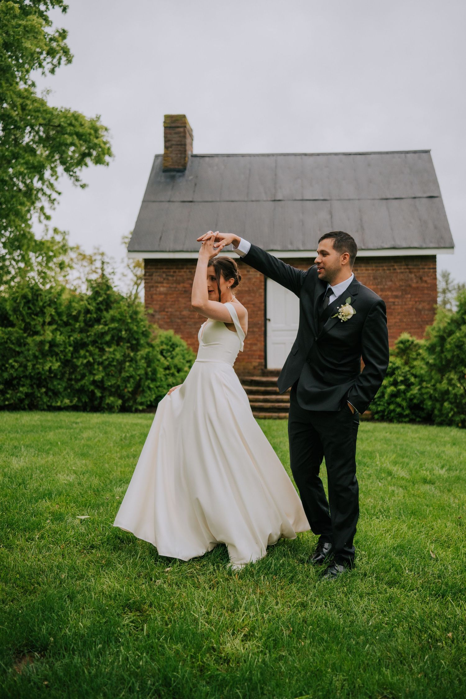 Couple dancing playfully on lawn with brick building in background during wedding celebration.