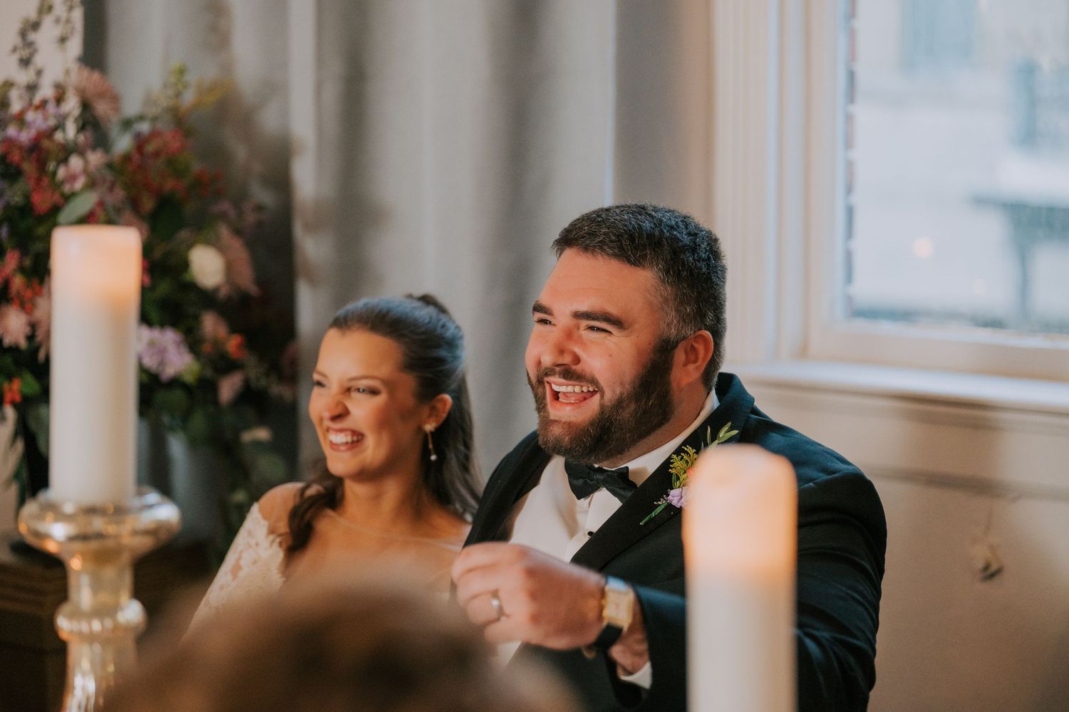 Wedding couple sharing joyful laughter during reception dinner with candlelight and floral decor.