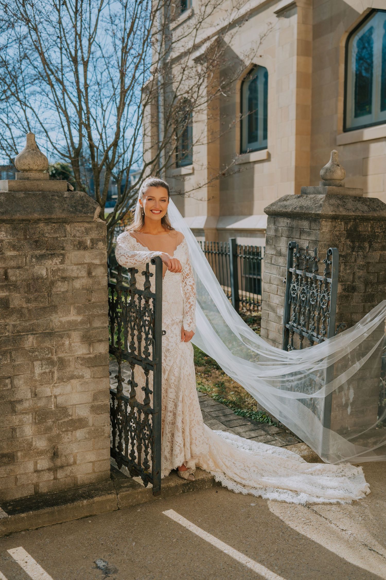 A beautiful bride in a lace wedding dress with flowing veil stands by an ornate iron gate outside a stone church.