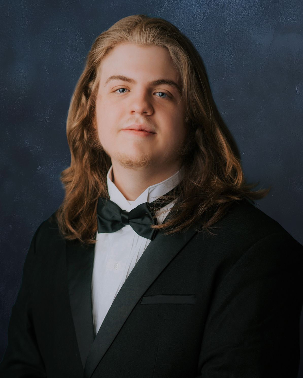 Young man with long brown hair wearing a black tuxedo and bow tie poses for a formal portrait photo.