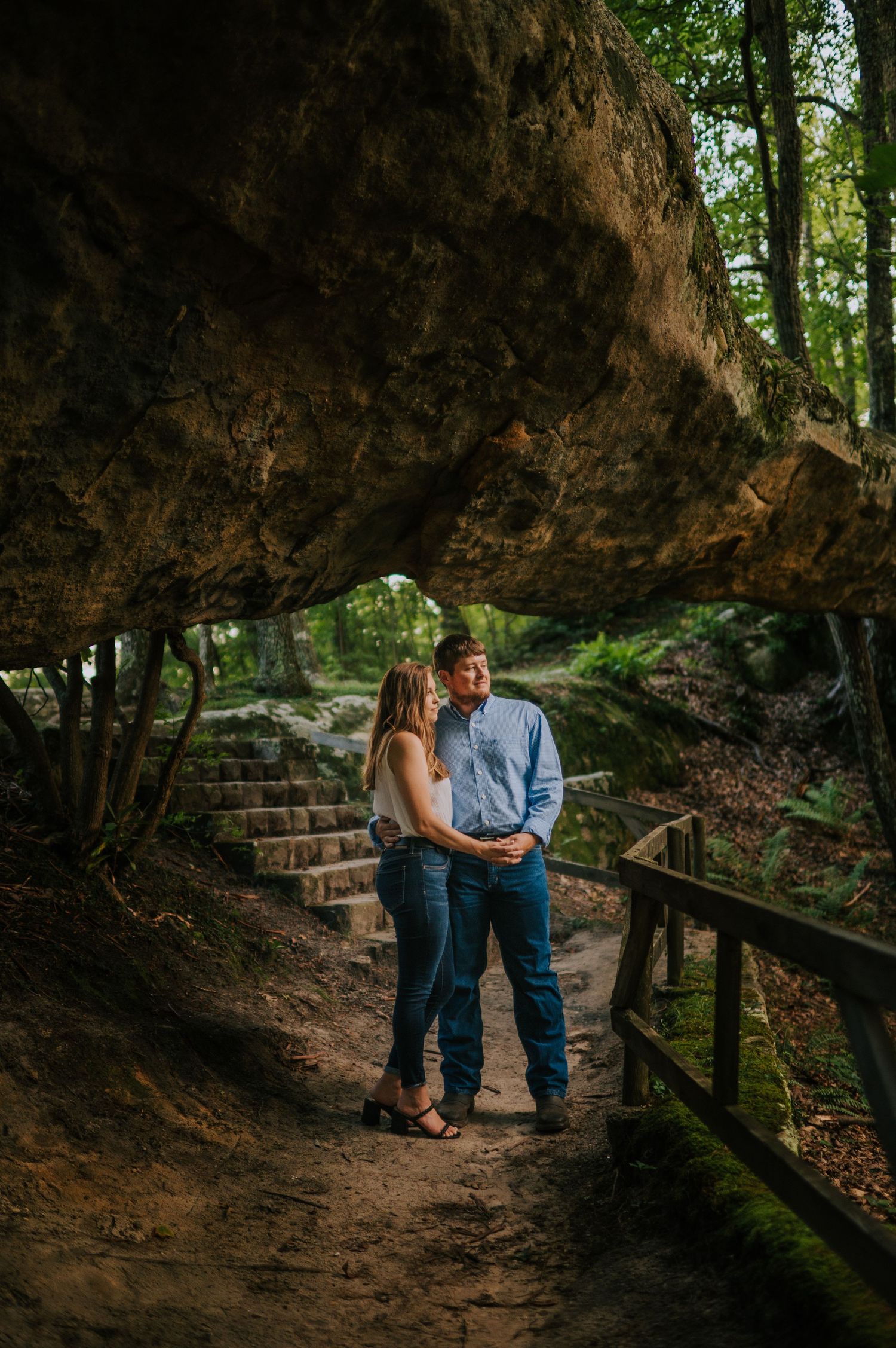 Romantic couple shares intimate moment under natural rock formation with stone steps in wooded setting.