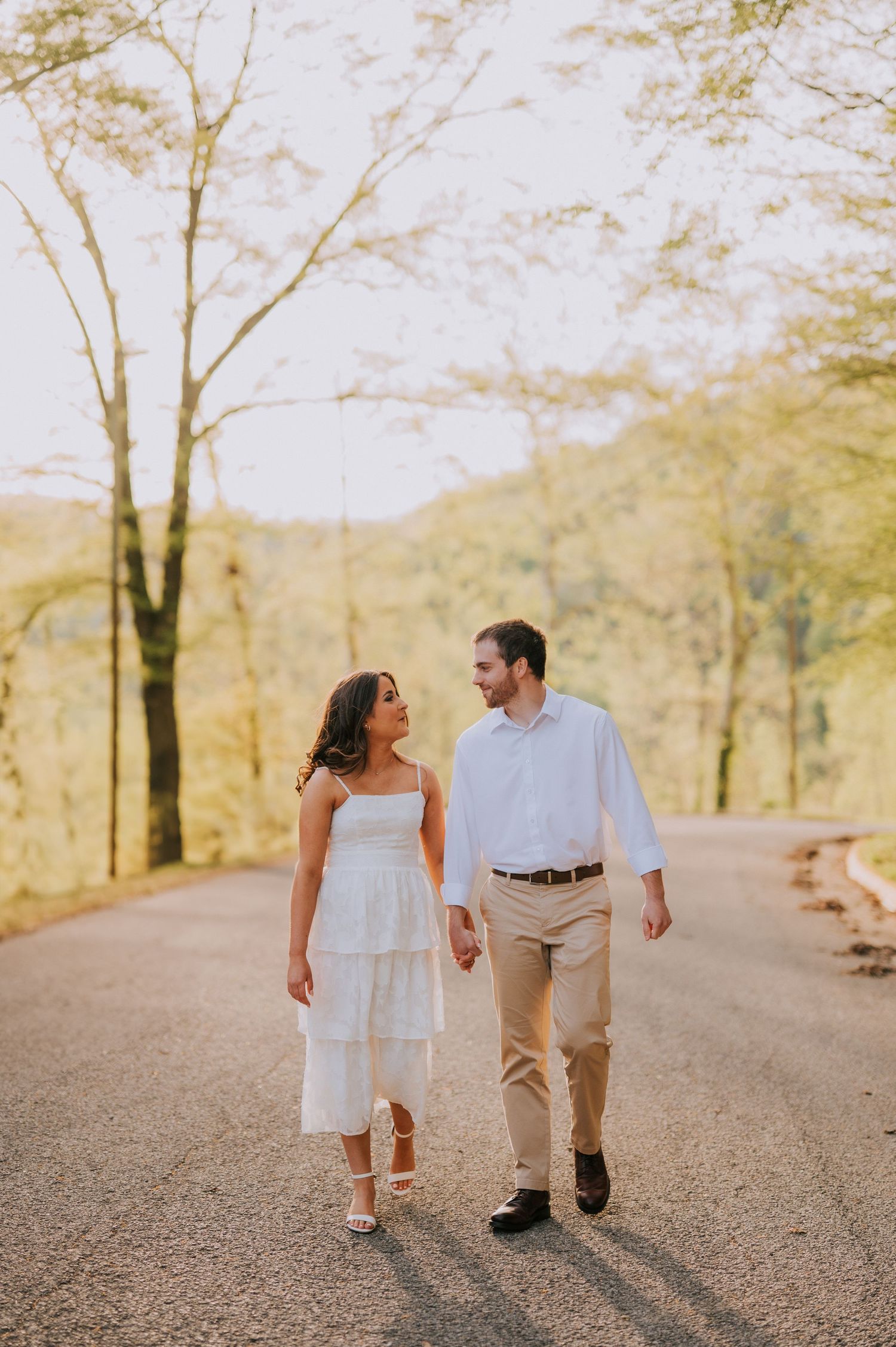 A couple walks hand in hand down a winding path surrounded by trees in warm sunlight.