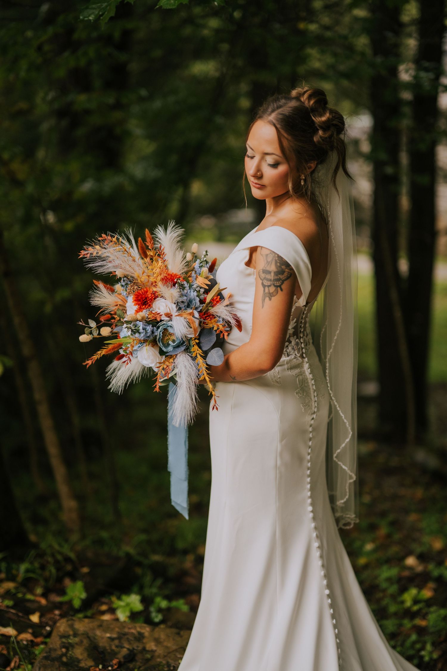 A wedding dress and bouquet featuring feathers and colorful flowers in a forest setting.