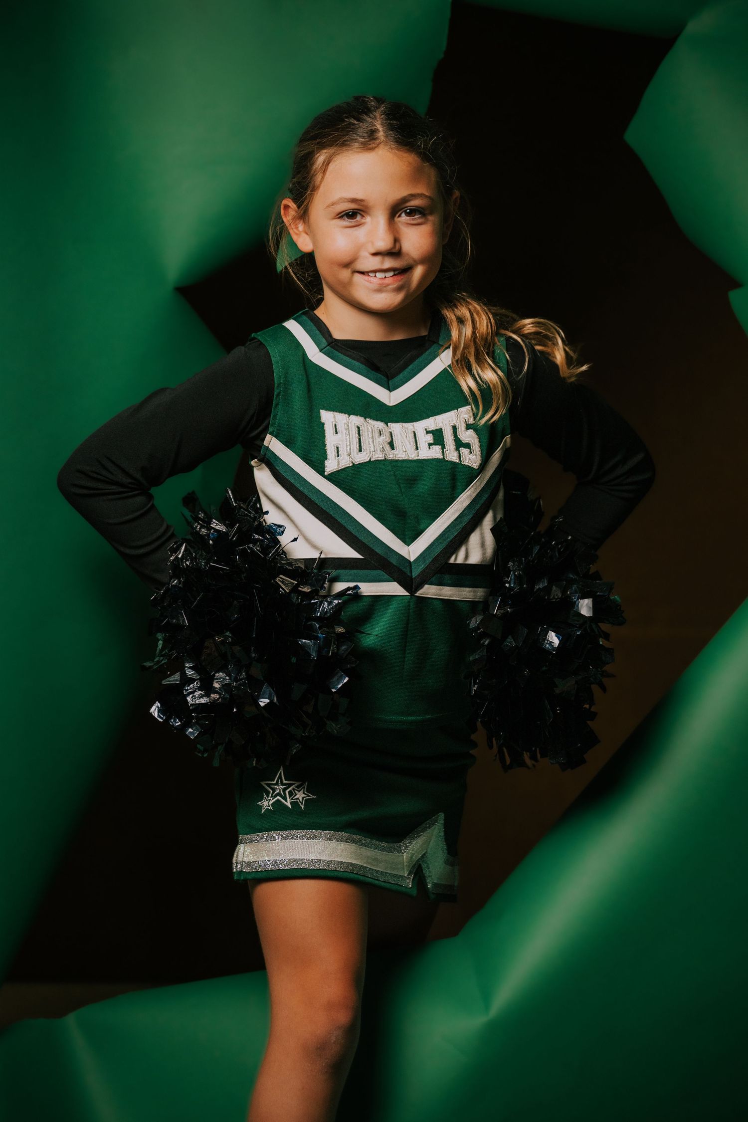 Cheerleader in green and white uniform with pom poms poses against dark background.