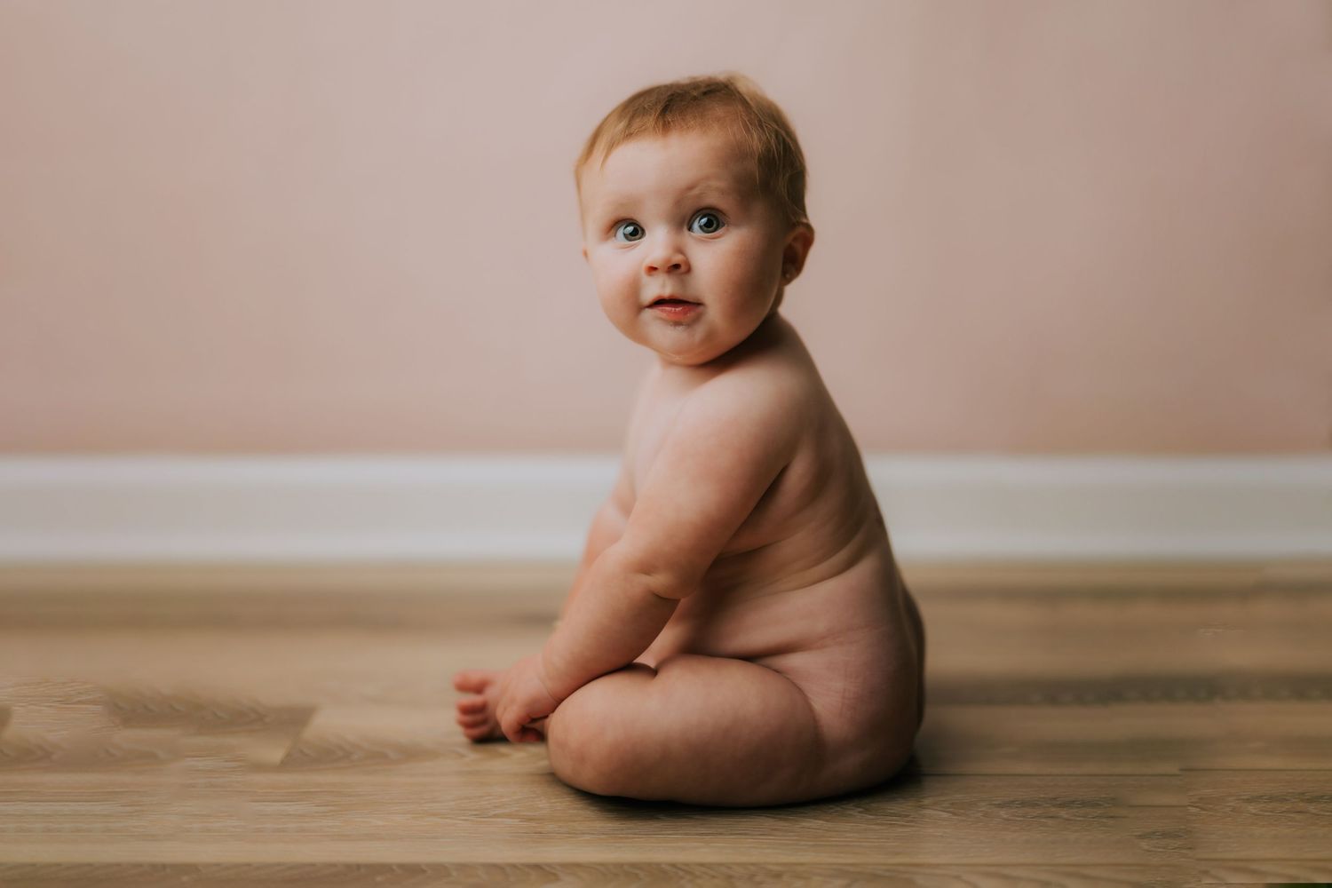Adorable baby sitting on hardwood floor against pink wall, looking over shoulder in a cute candid portrait photo.