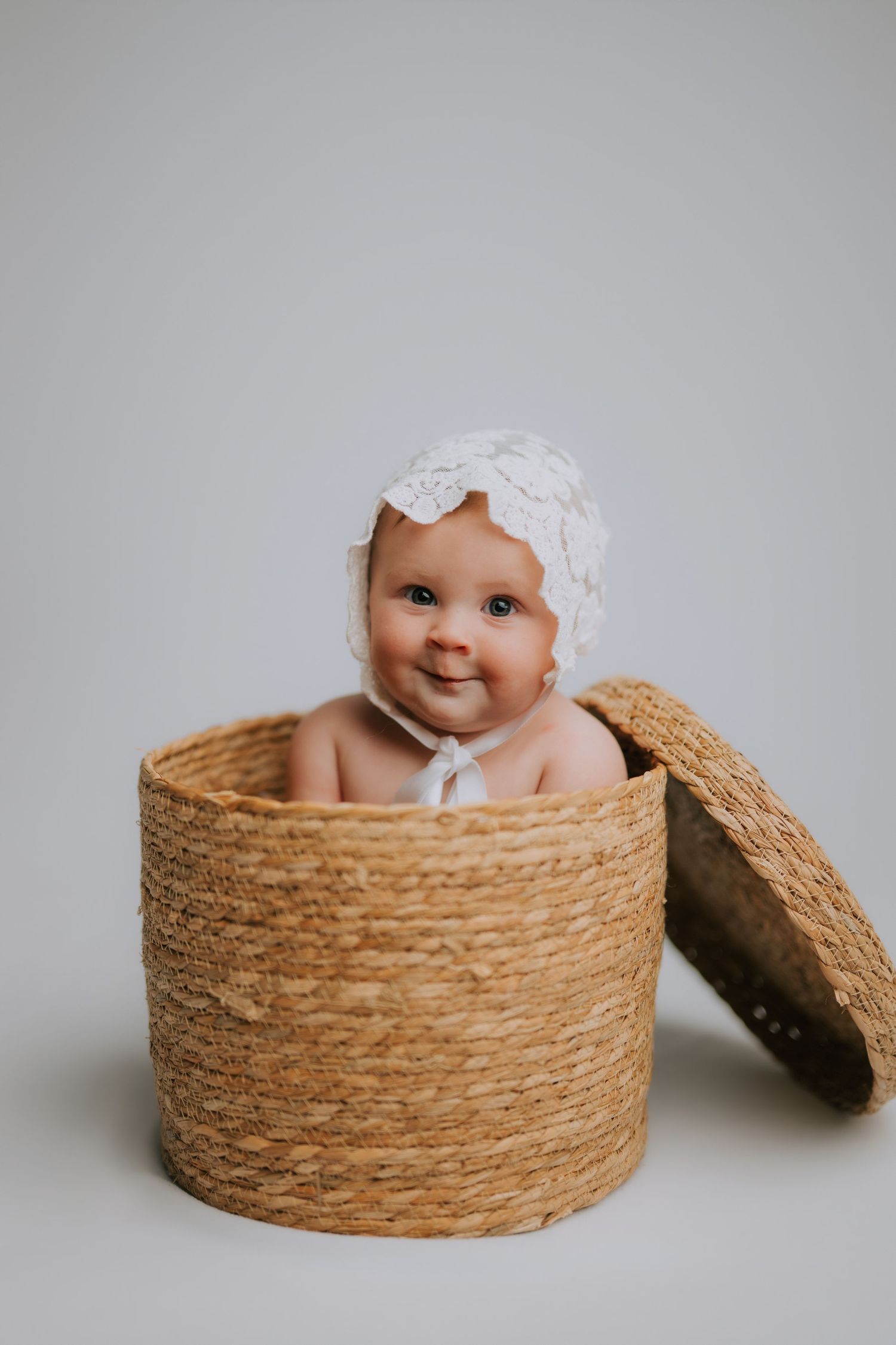 Smiling baby wearing a white lace bonnet sitting inside a woven wicker basket on a light gray background.