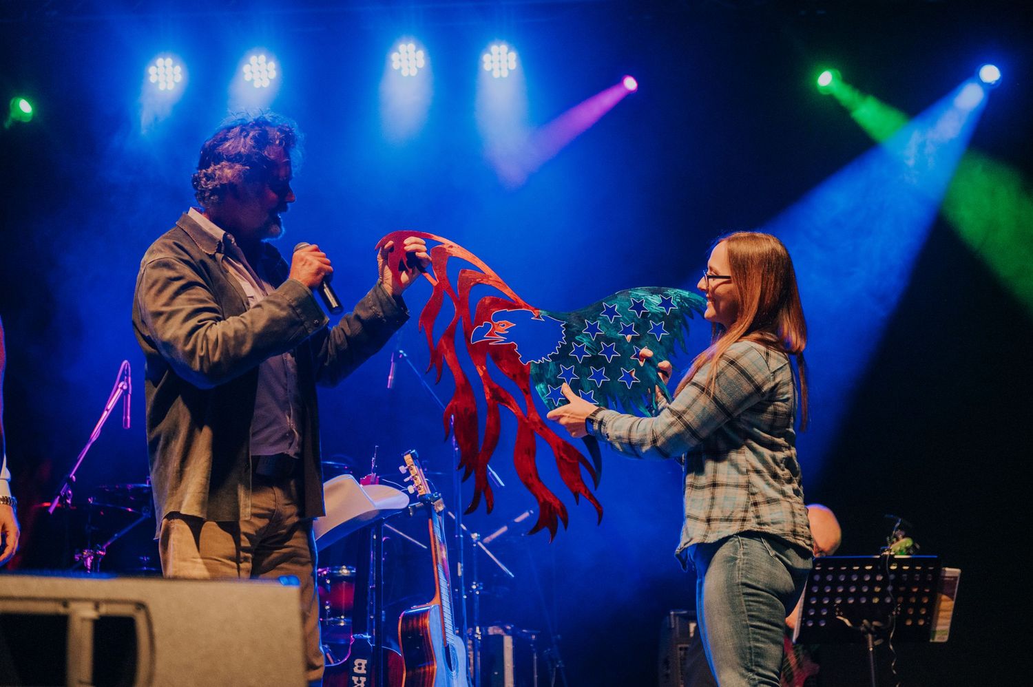 Two performers interacting on stage with colorful blue and green spotlights in the background.