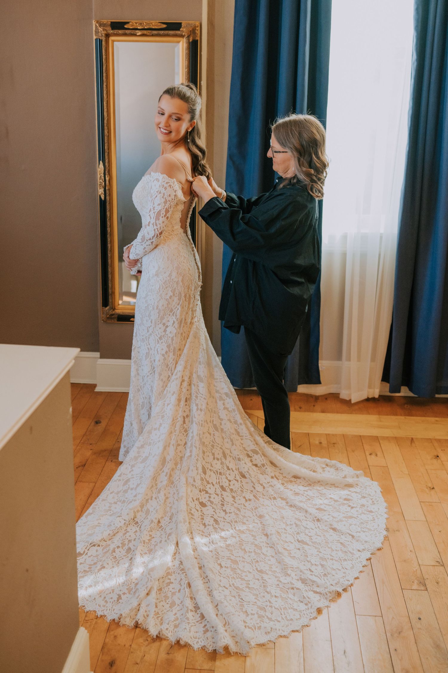 A bride wearing a stunning lace wedding dress with a long train gets help with final adjustments in front of a mirror.