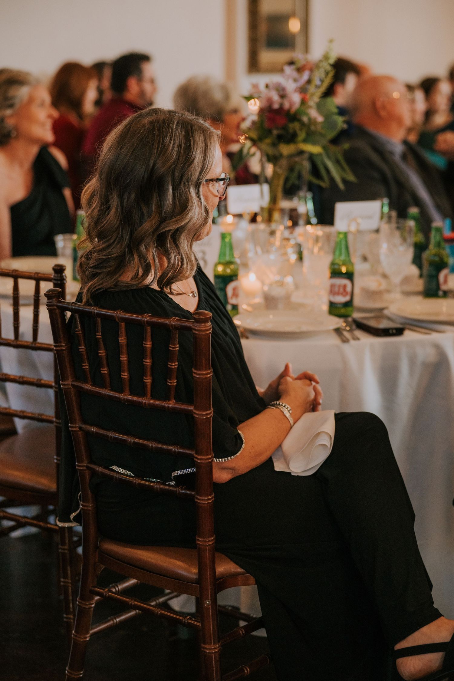 Event attendee in black dress seated at elegantly decorated reception table with floral centerpiece and bottles.