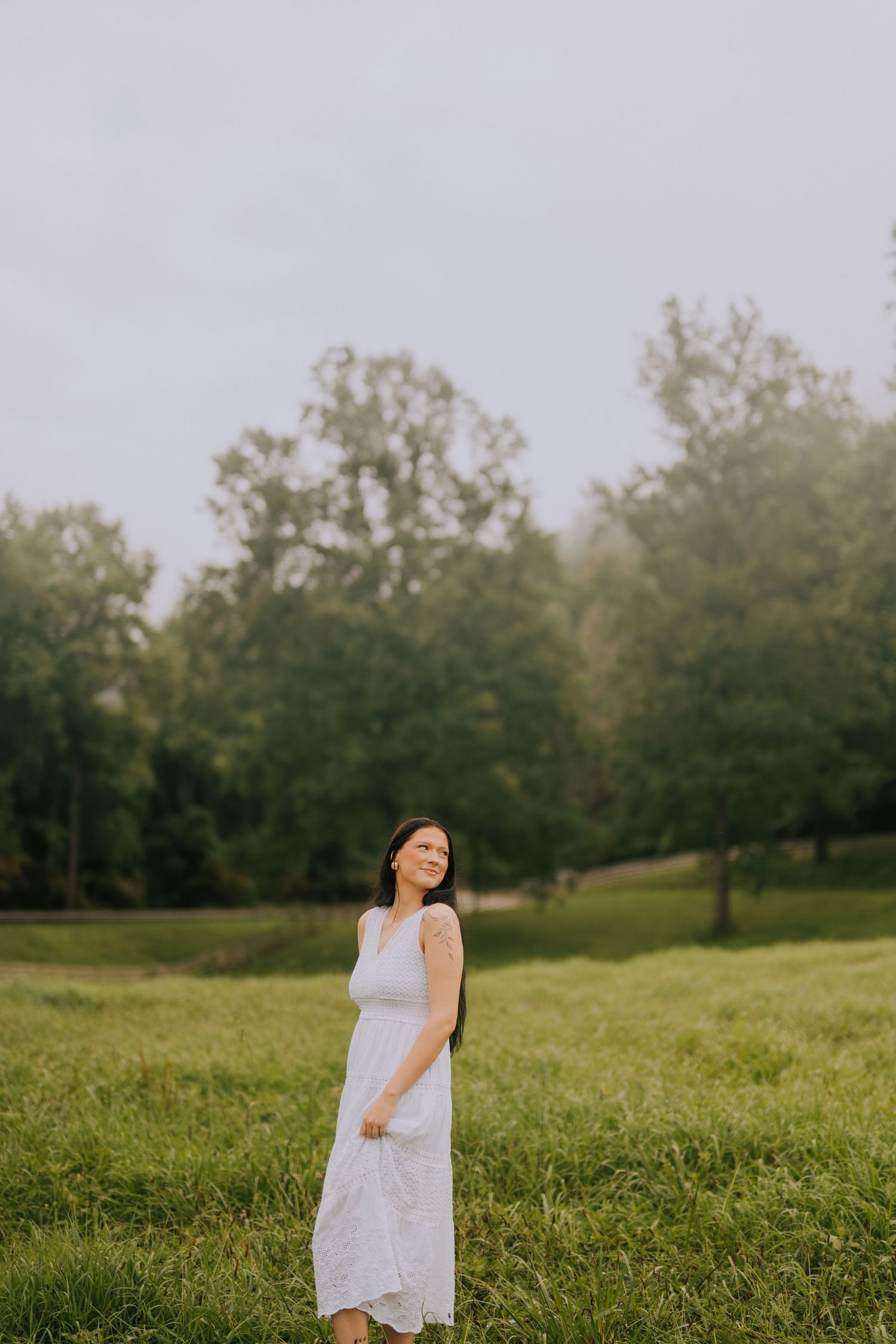 Person in white dress poses gracefully in open field during overcast day.