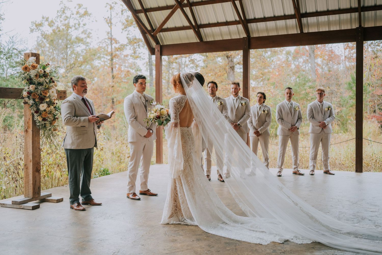 Wedding party stands under a covered pavilion during an autumn outdoor ceremony.
