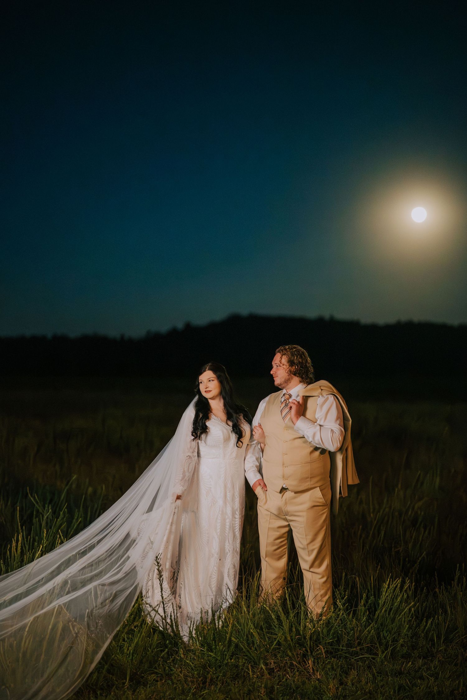 A romantic wedding couple poses in a field under a glowing full moon at dusk.