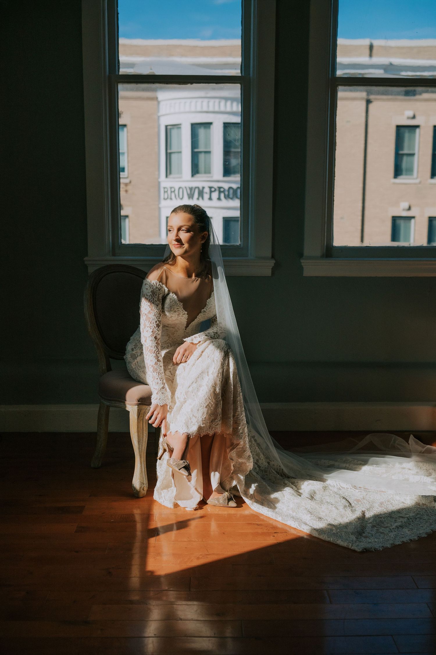 Bride in vintage-style lace gown sitting in sunlit room with flowing veil and city view.