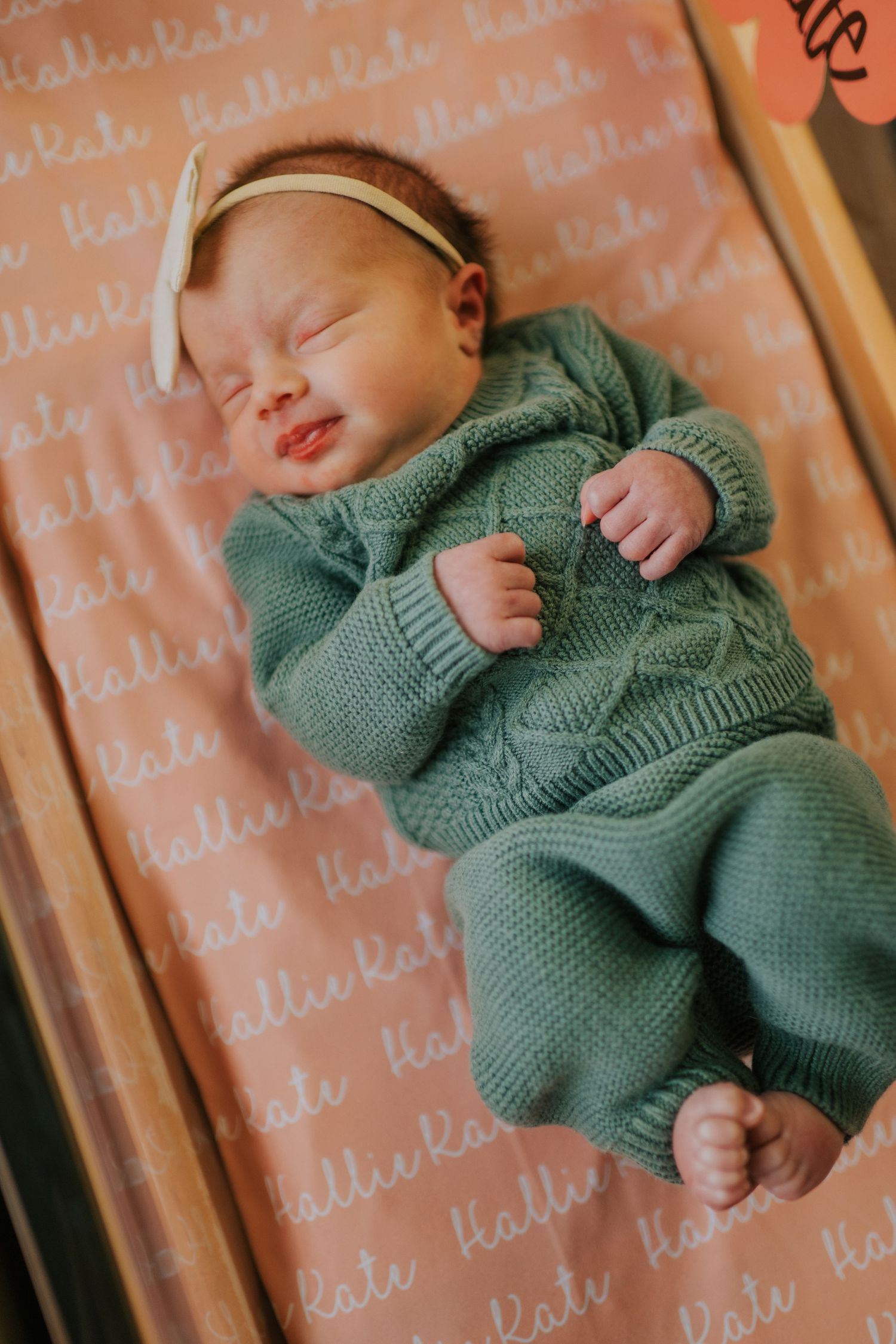 A newborn wearing a sage green knit sweater rests peacefully on a textured surface.