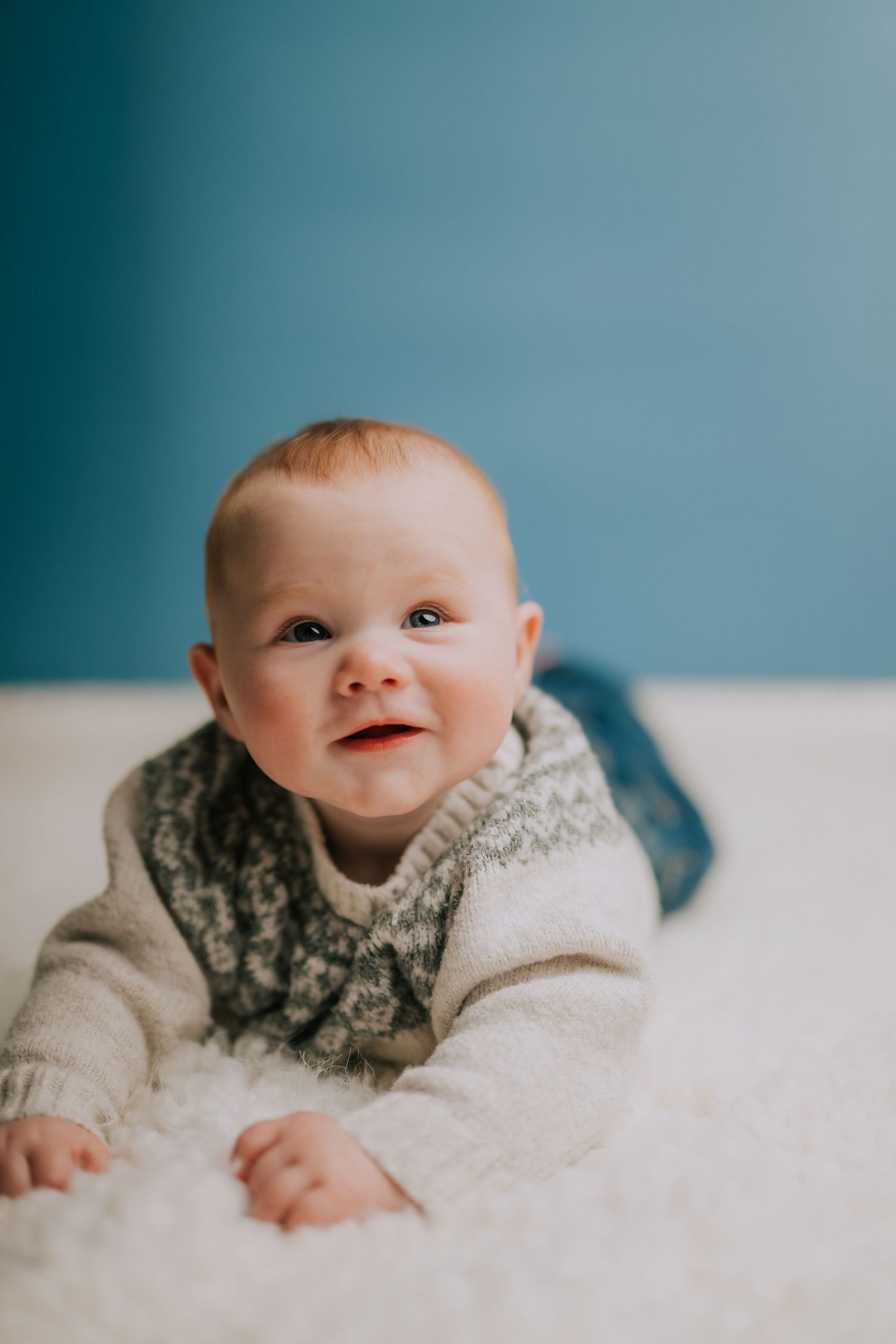 Baby in knit sweater lying on soft white blanket against blue background.