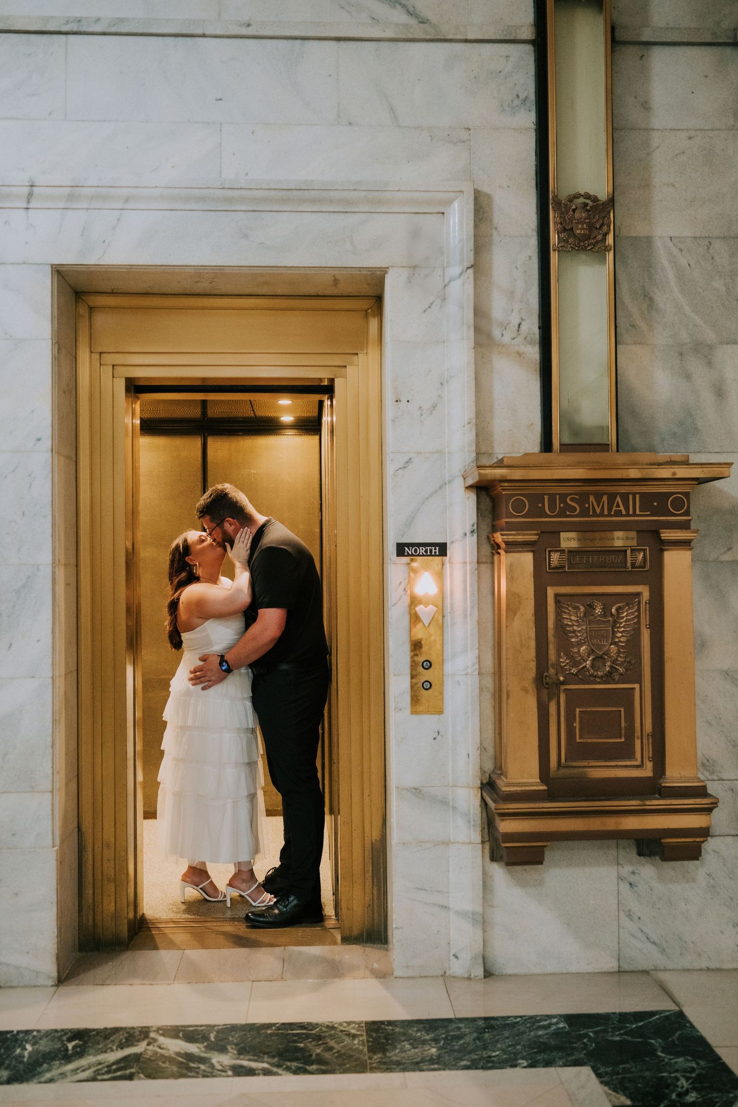 A couple embraces near vintage mailboxes in a marble-walled hallway.