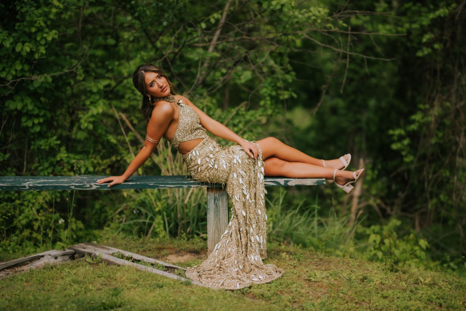 Woman in gold sequin gown poses elegantly on a bench surrounded by lush green trees.