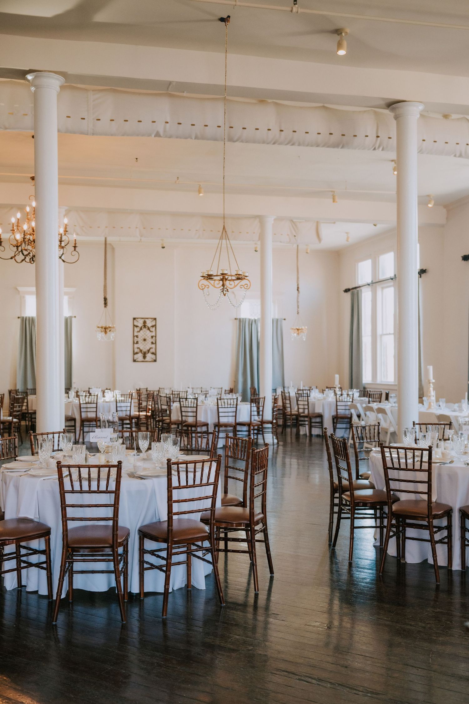 Elegant banquet hall with white columns, round tables with white linens, and wooden Chiavari chairs set for a formal event.