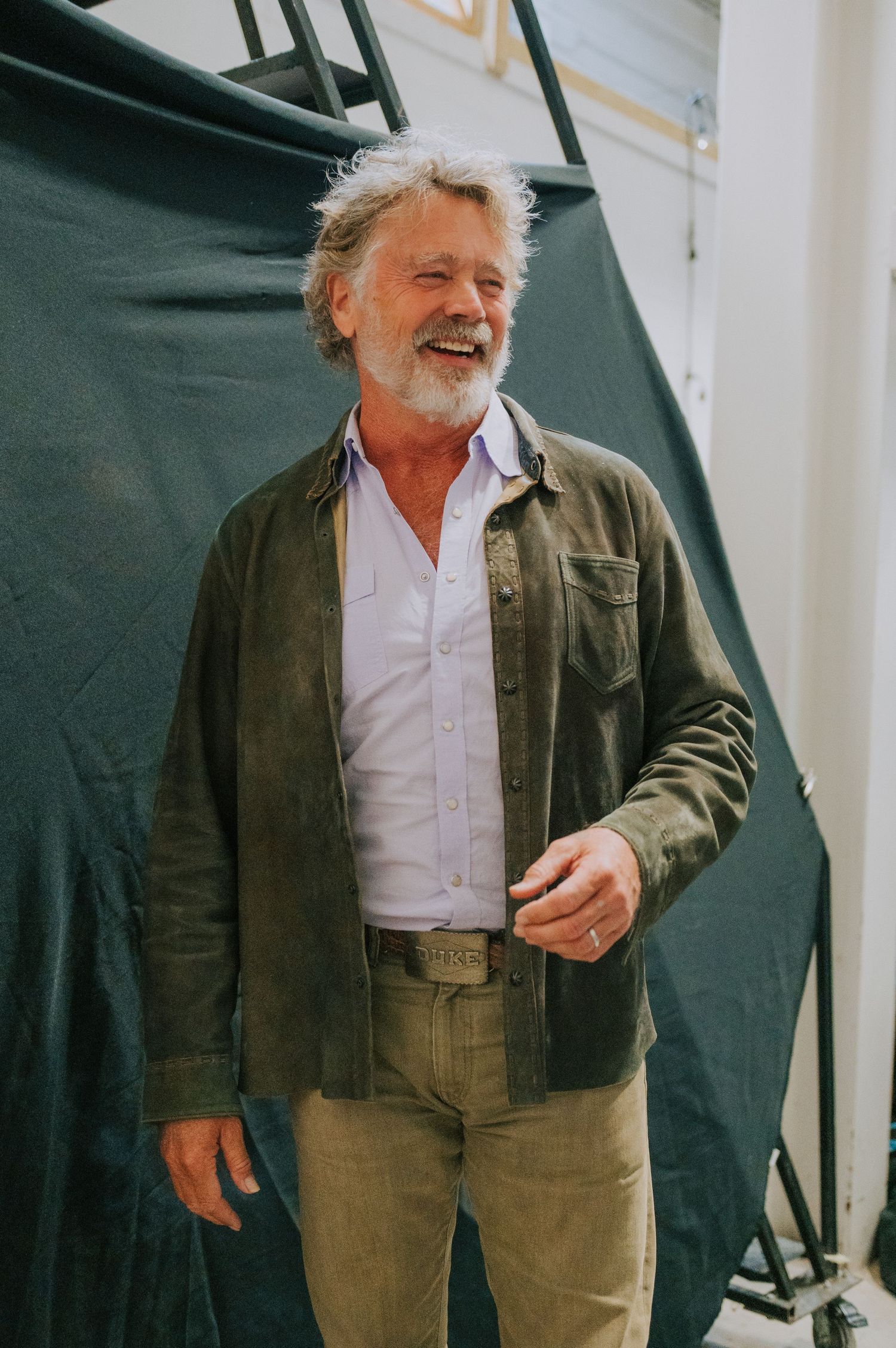 A person in a green jacket and khakis stands outdoors smiling warmly against a dark green background.