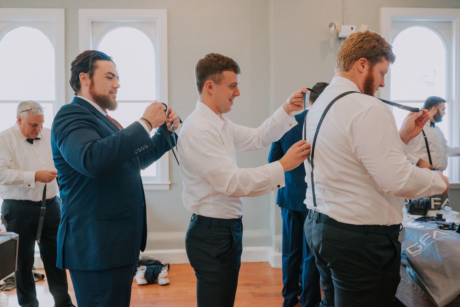 Groomsmen getting ready in a bright room with arched windows adjust their suspenders and ties before a wedding celebration.