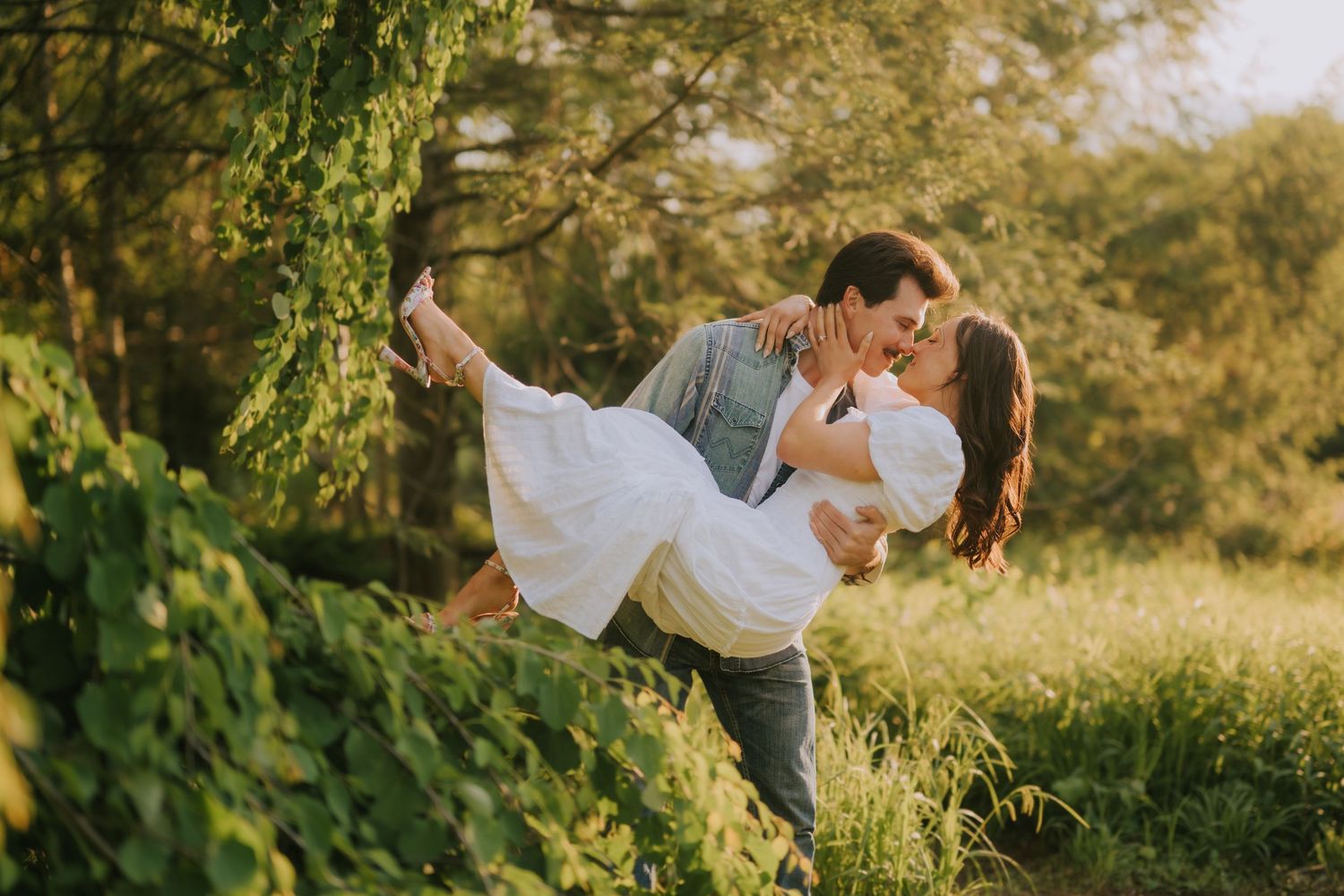 Romantic couple sharing a kiss outdoors, man lifting woman in white dress among lush green trees in golden sunlight.