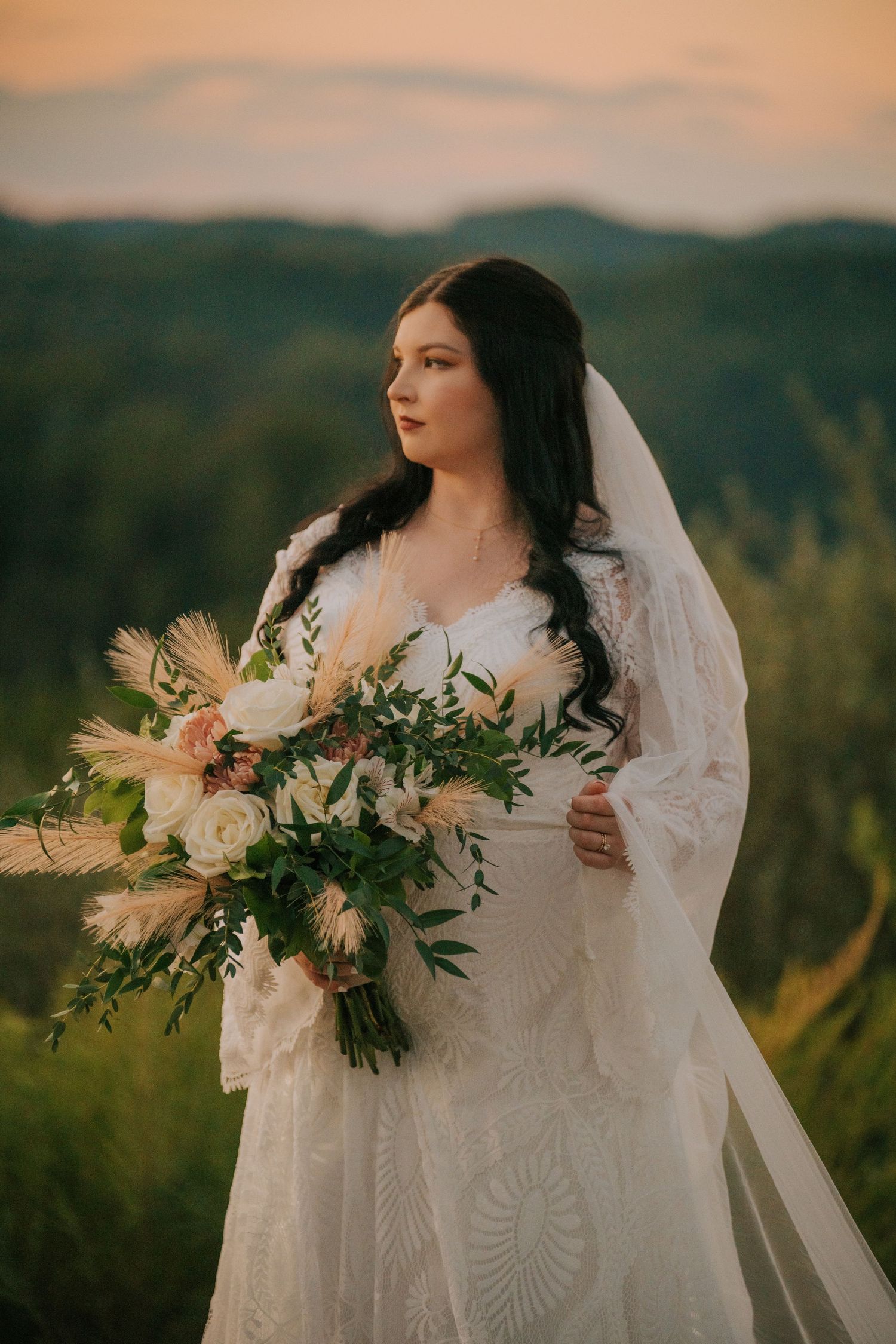 Bride in lace gown holding natural bouquet against mountain landscape at sunset.