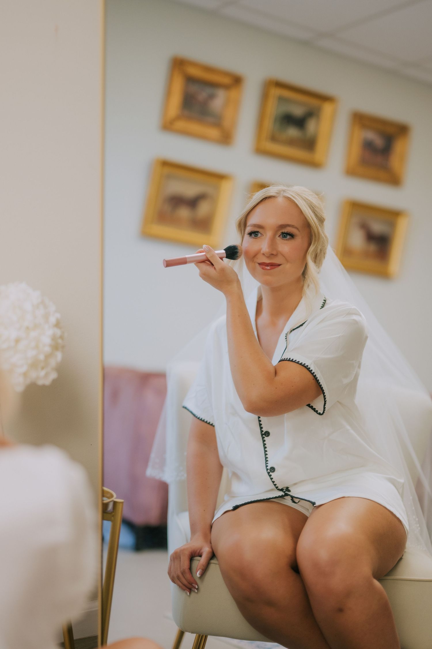 Getting ready scene with bridal preparations in white robe against wall with framed artwork.