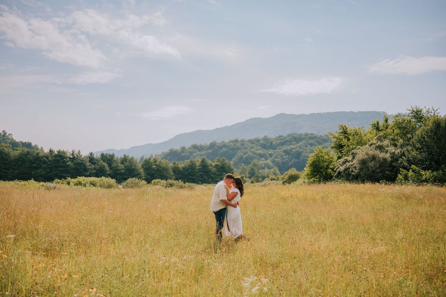 A couple embraces in a golden meadow with rolling mountains and forest in the background.