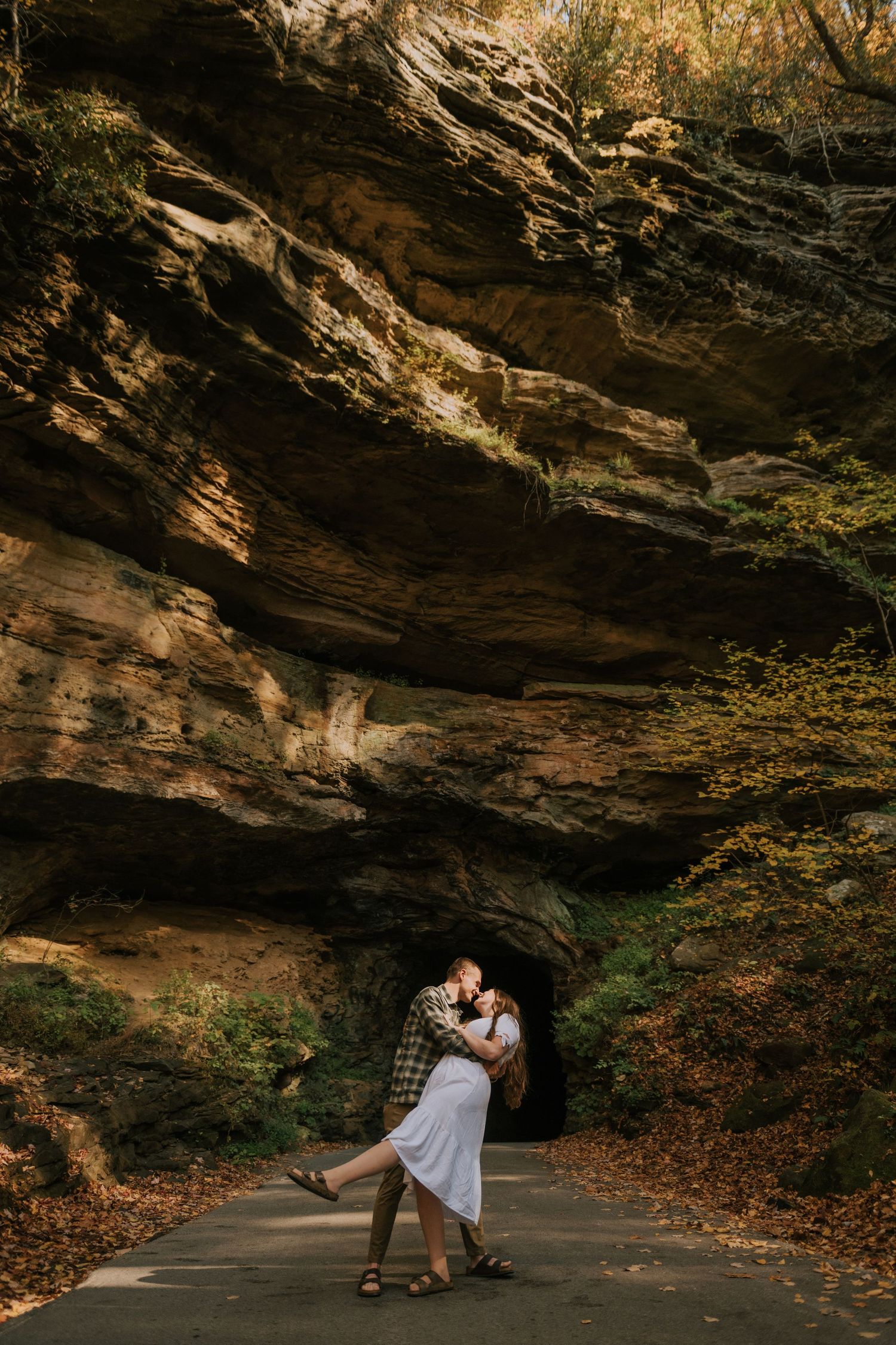 A romantic couple embraces beneath a dramatic rock overhang in an autumn forest setting.