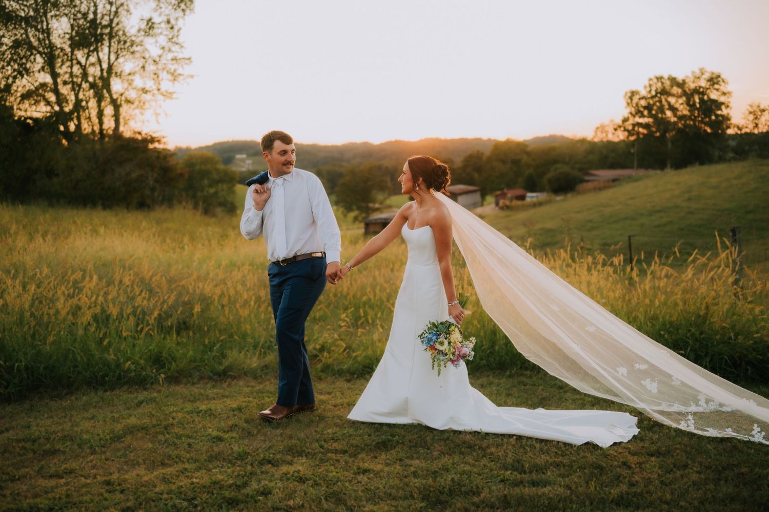 Wedding couple poses in golden field with flowing veil catching sunset light.