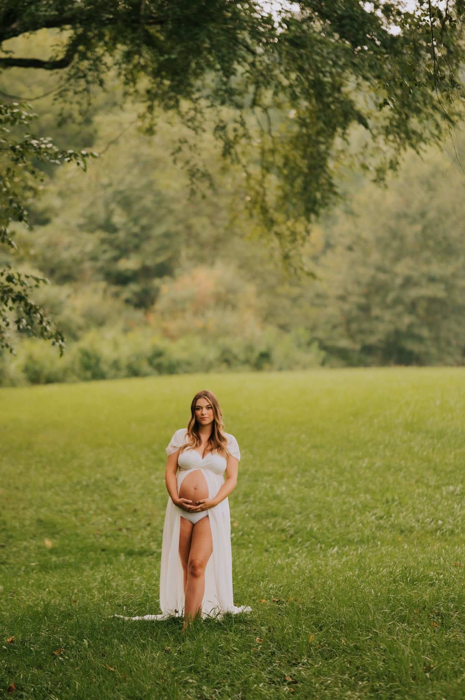 A maternity photoshoot in a sunny meadow with a woman in a flowing white dress surrounded by trees.