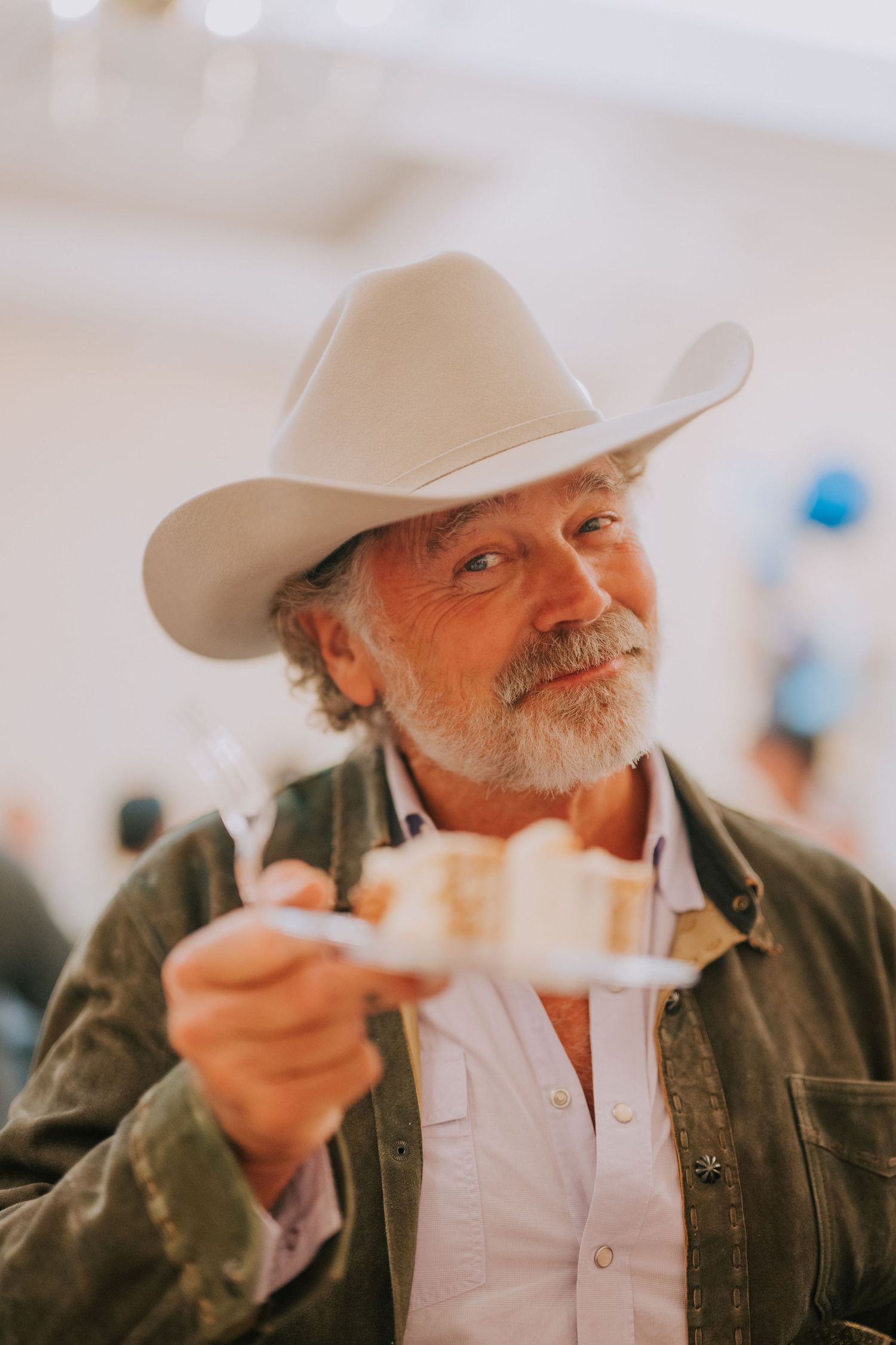 A mature man wearing a cowboy hat and jacket smiles while holding something in his hand.