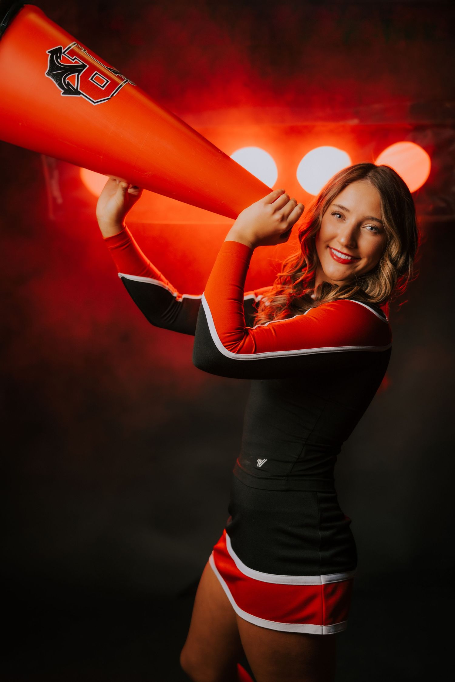 Cheerleader in black and red uniform poses with orange megaphone against dark background with glowing red lights.