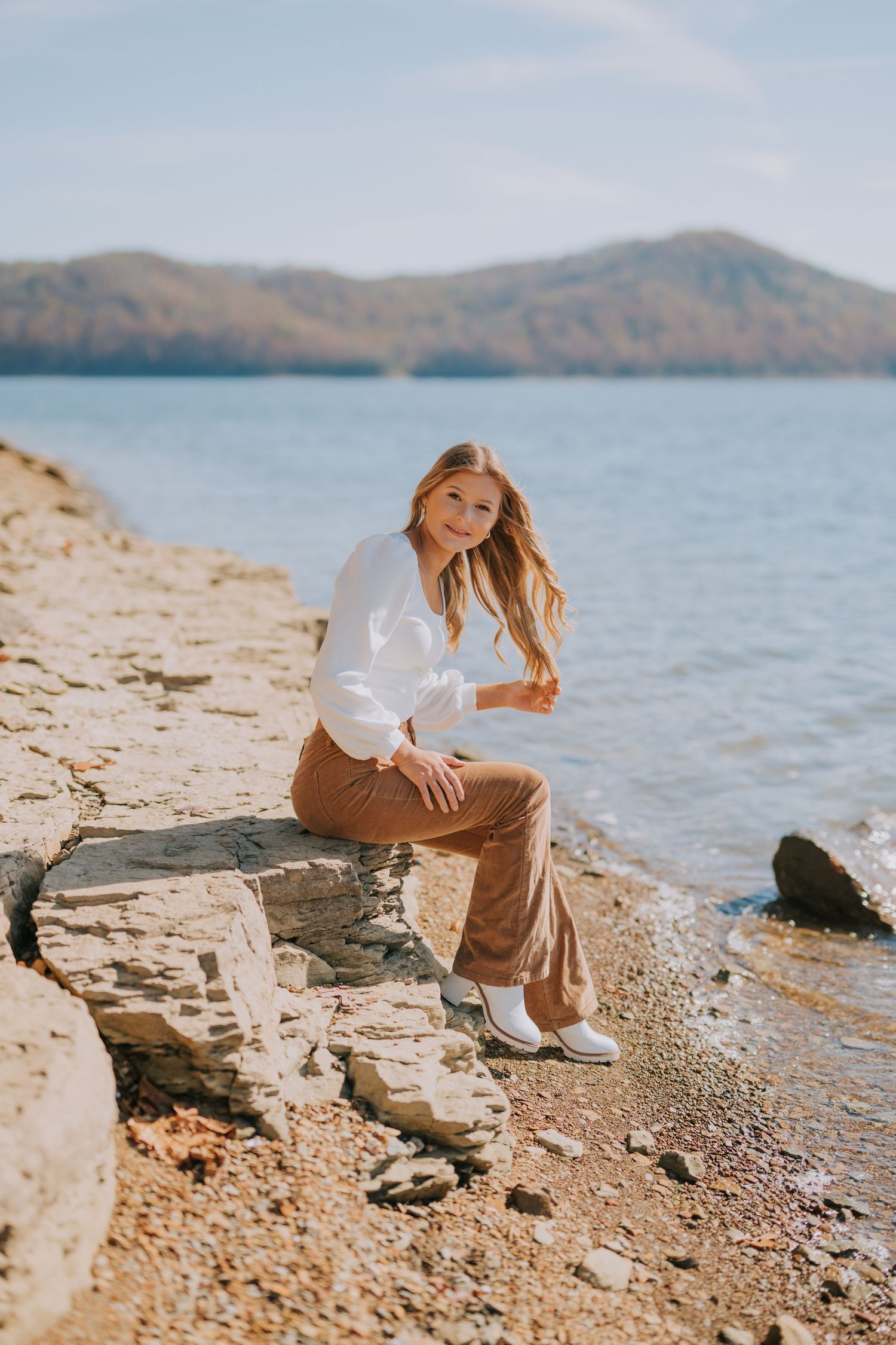 Person sitting on rocky shoreline with mountains and lake in background during golden hour.