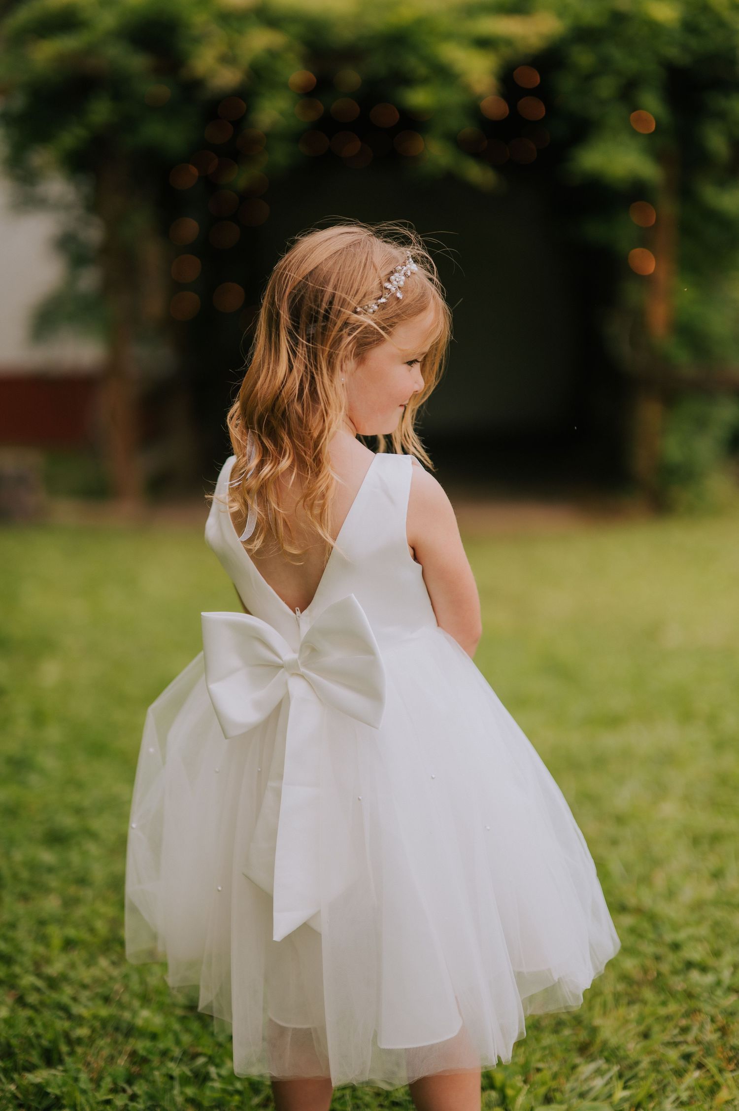 Little flower girls in white tulle dresses with large bows twirl on the grass.