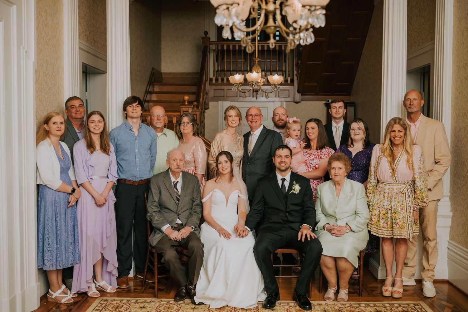 Large wedding party group photo featuring guests in pastel dresses and formal attire gathered in an elegant interior.