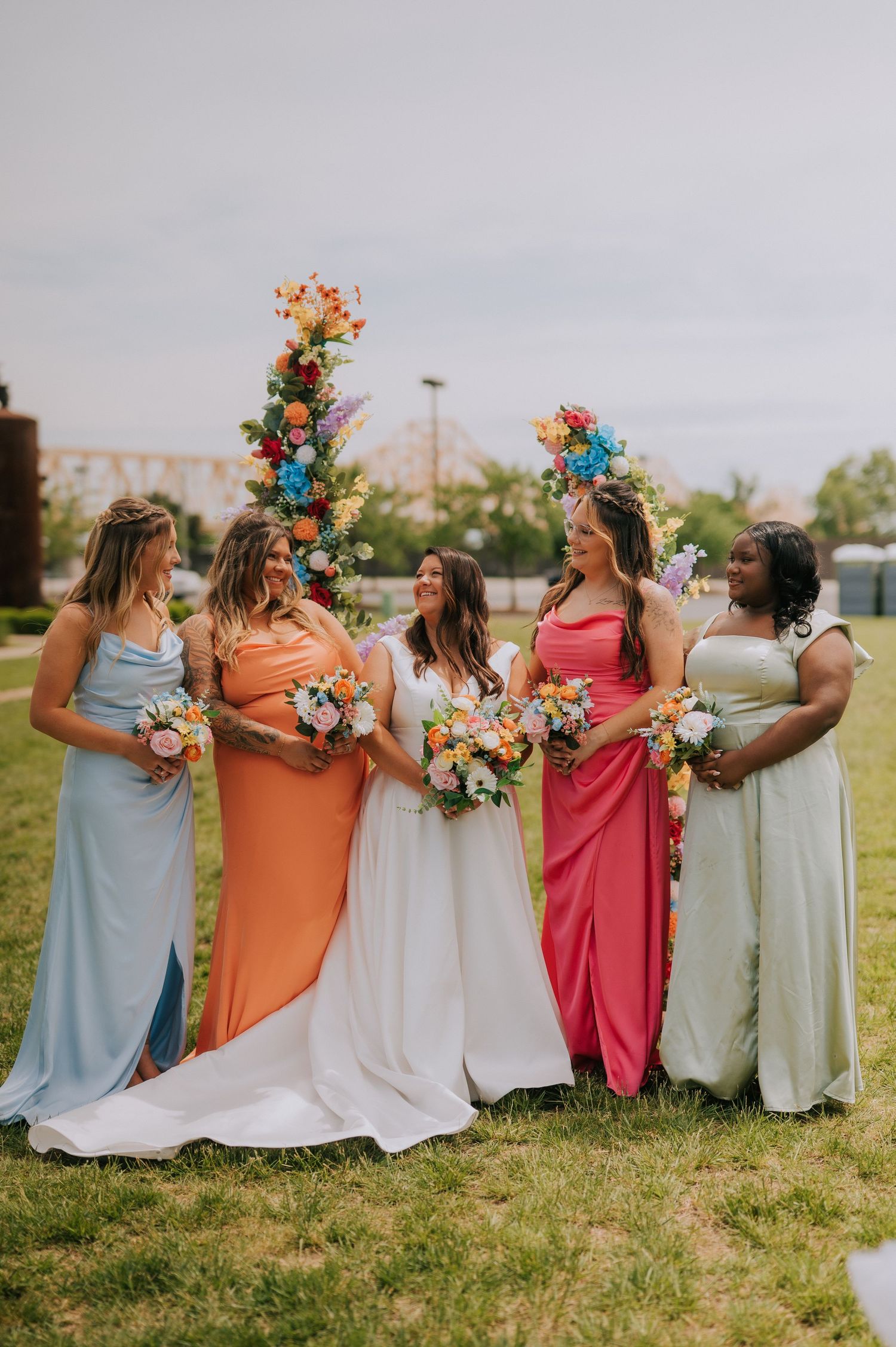 Bridesmaids in pastel dresses holding colorful bouquets pose together on grassy field.
