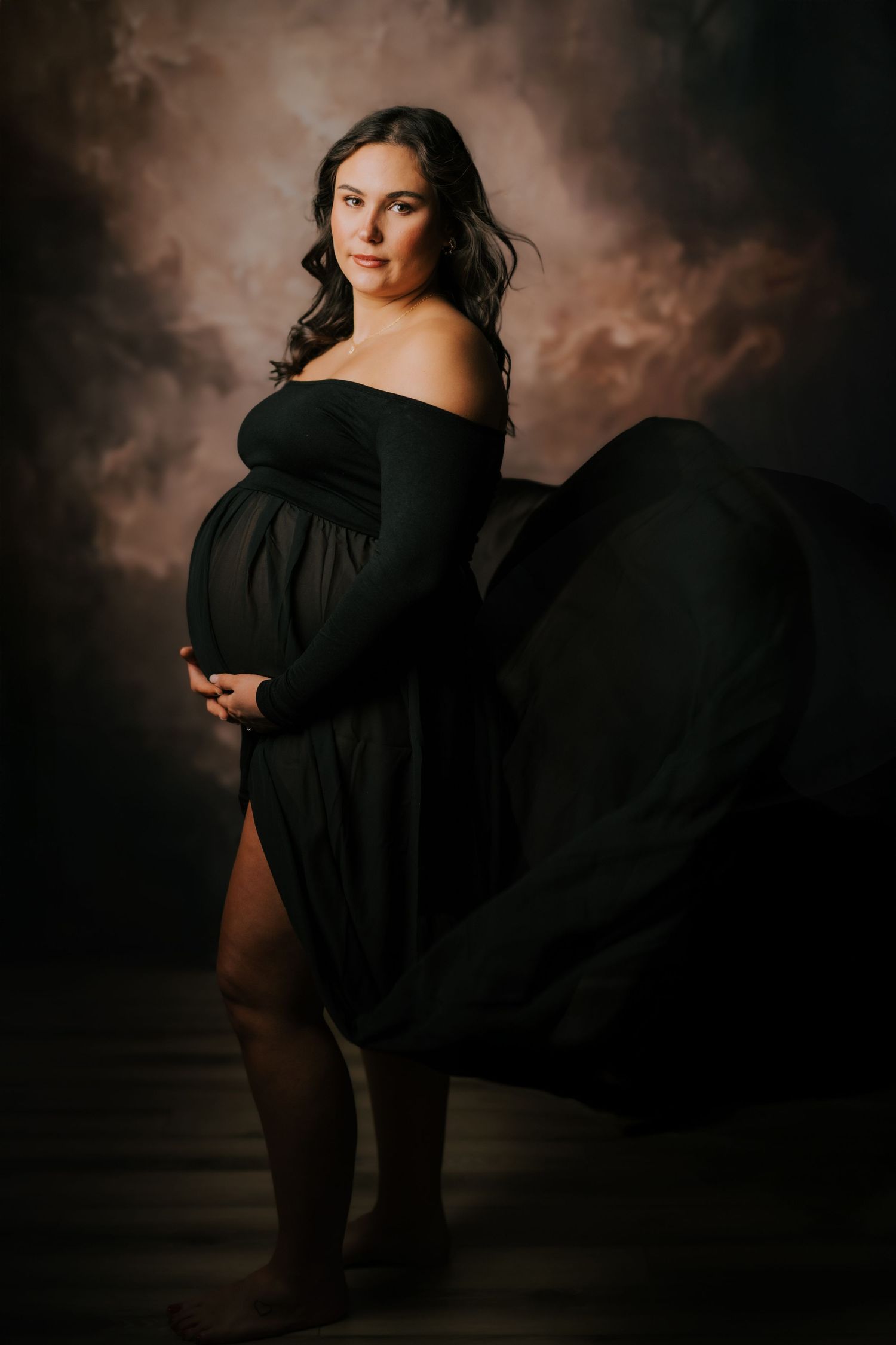 A dramatic maternity portrait features an elegant black off-shoulder gown flowing against a moody brown backdrop.