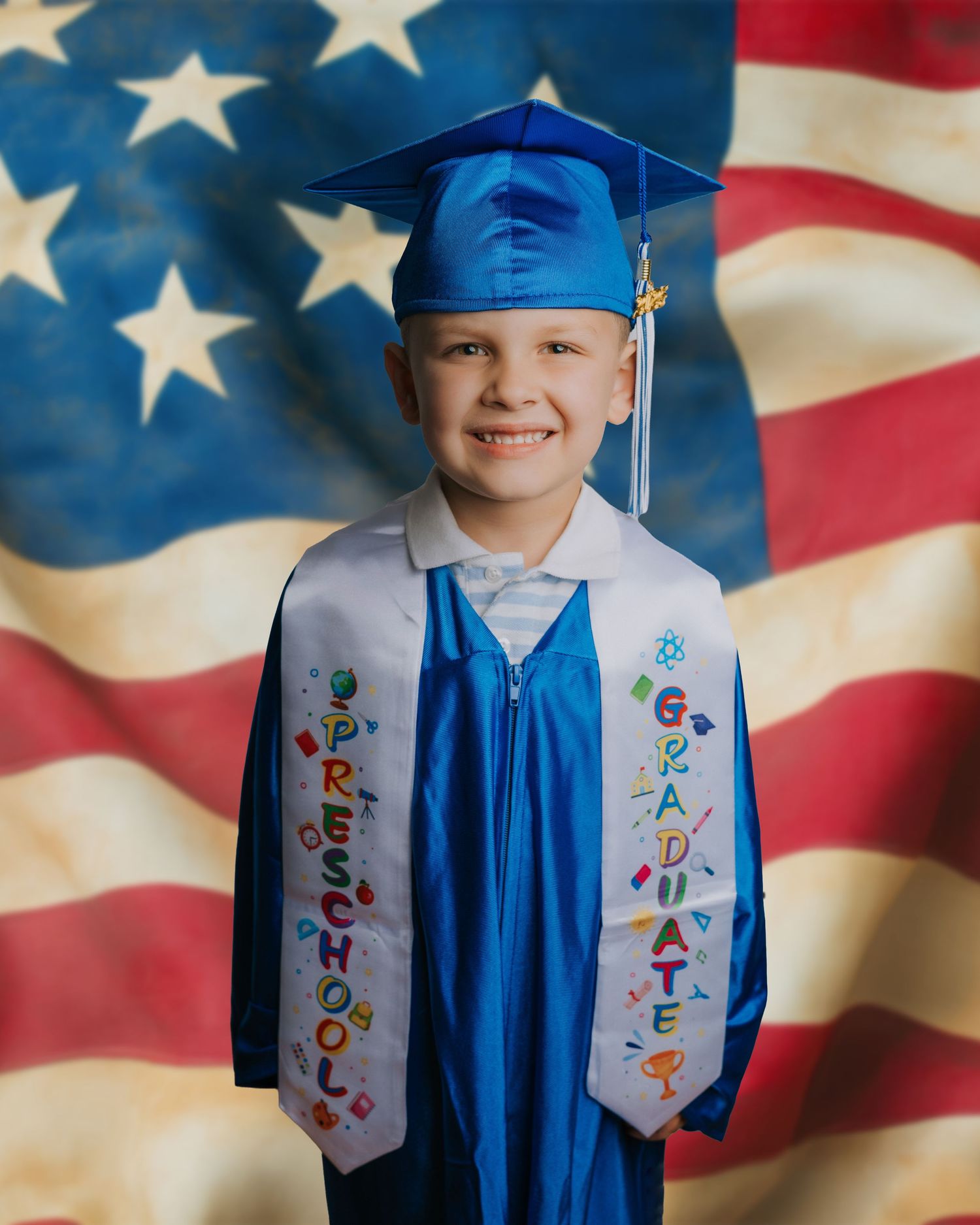Young child in blue cap and gown with preschool graduate sash smiling in front of American flag backdrop.