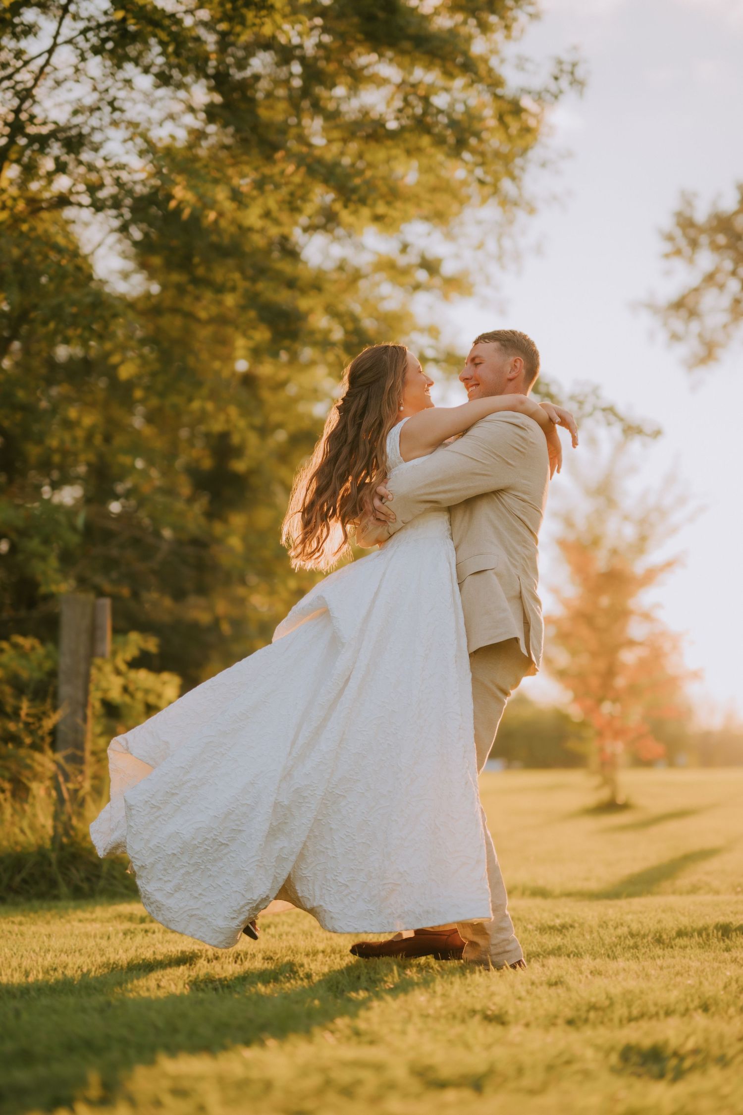 Groom lifts bride in white gown during golden hour outdoor wedding portrait surrounded by lush green trees.