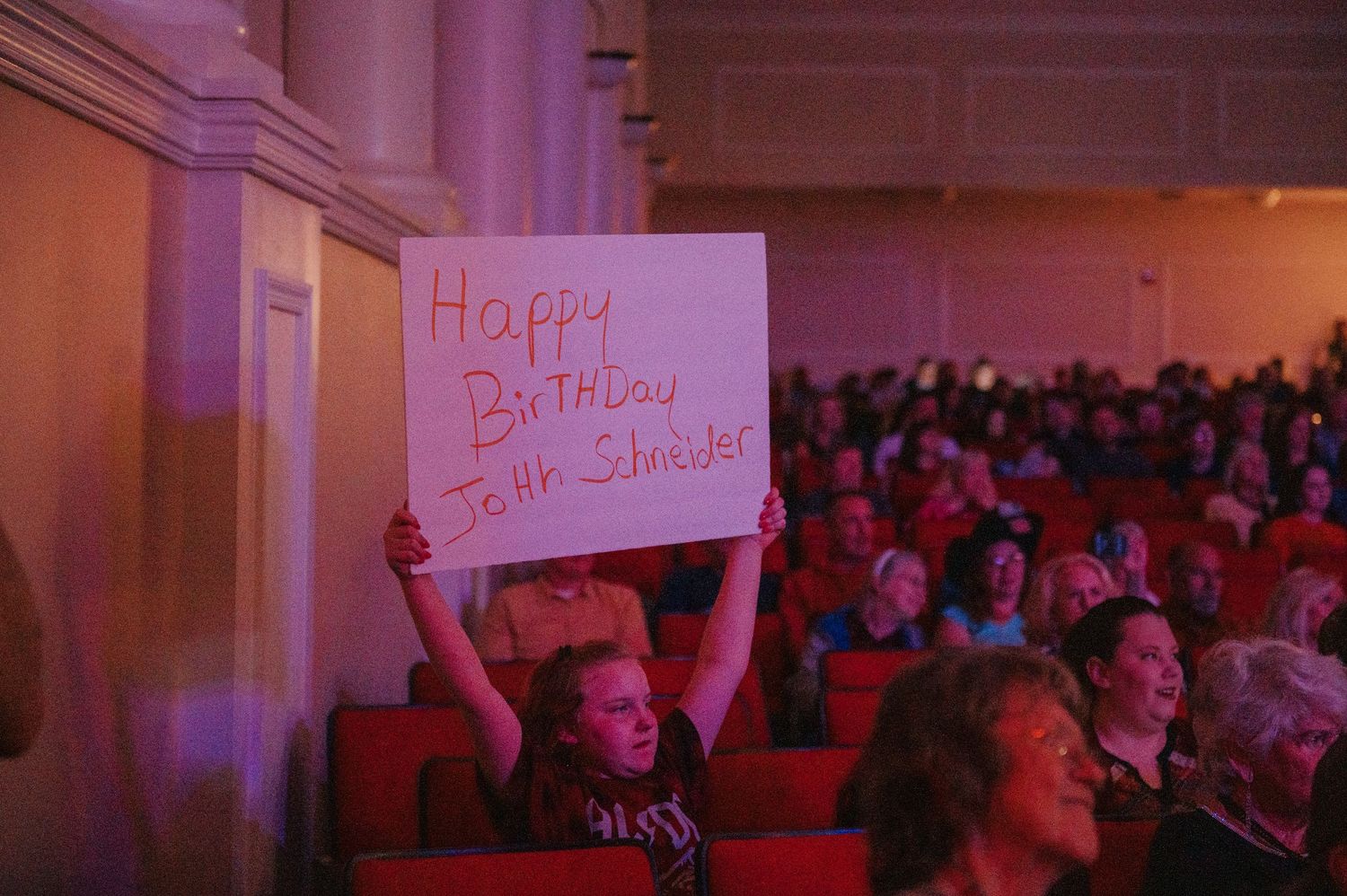 Concert audience member holds up 'Happy Birthday' sign during performance in a dimly lit venue with pink lighting.