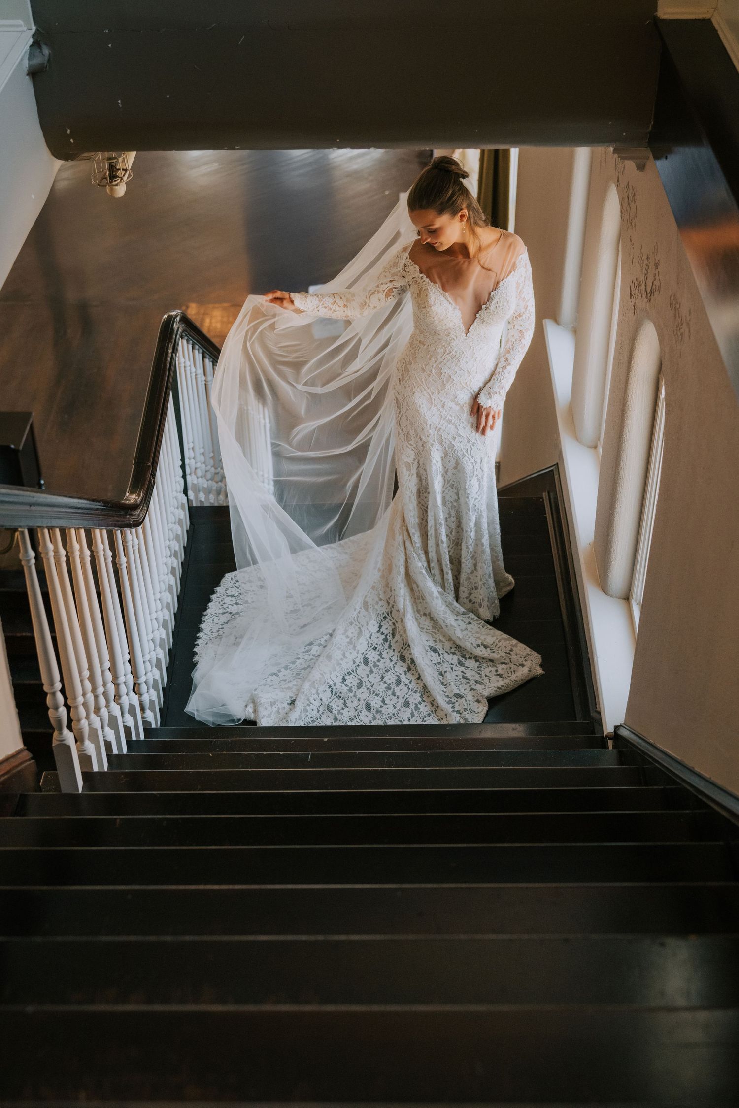 A bride in an elegant lace wedding gown descends a dark staircase while holding her flowing veil in dramatic lighting.