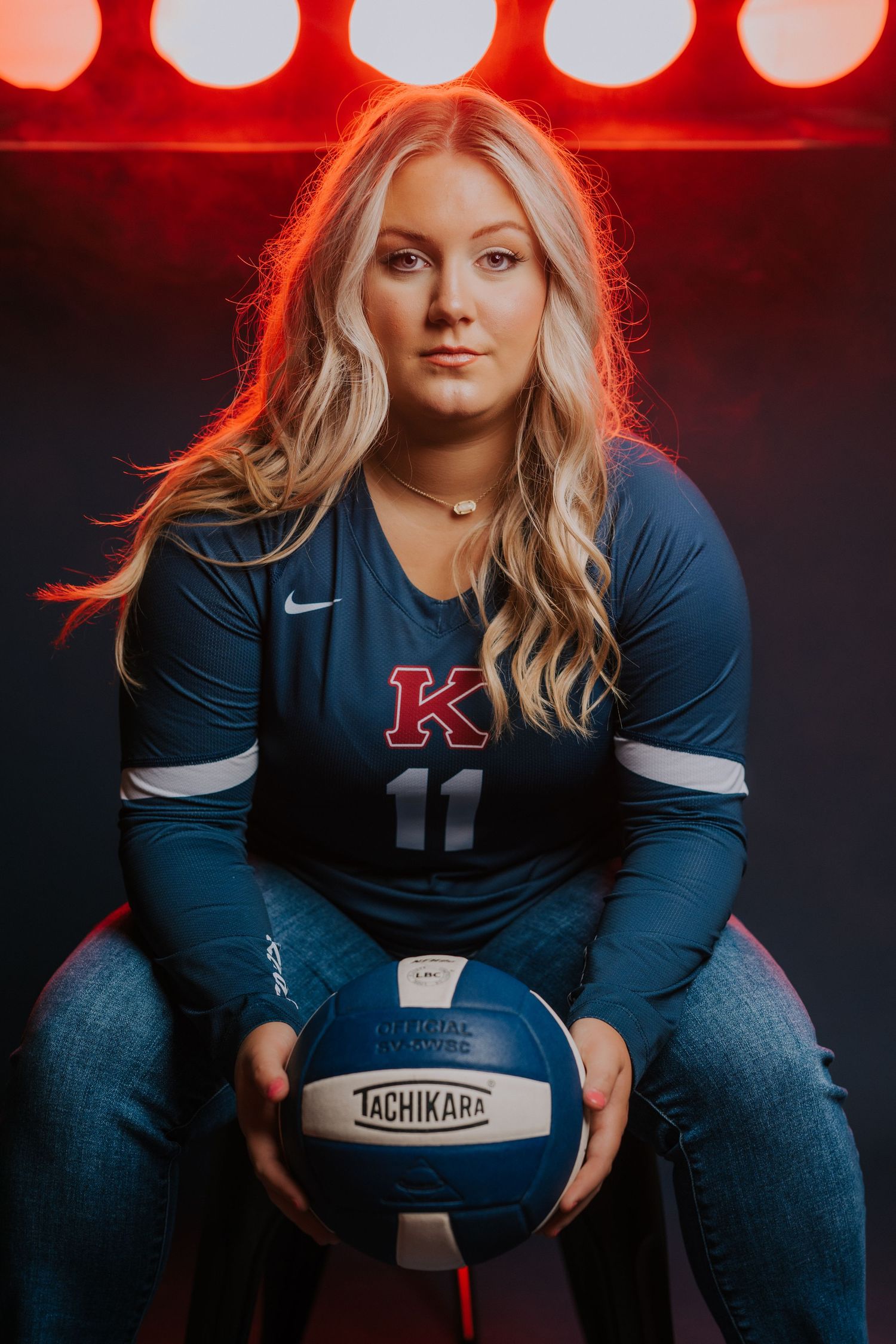 Athletic volleyball player in navy blue team jersey poses with ball against red backdrop with dramatic lighting.