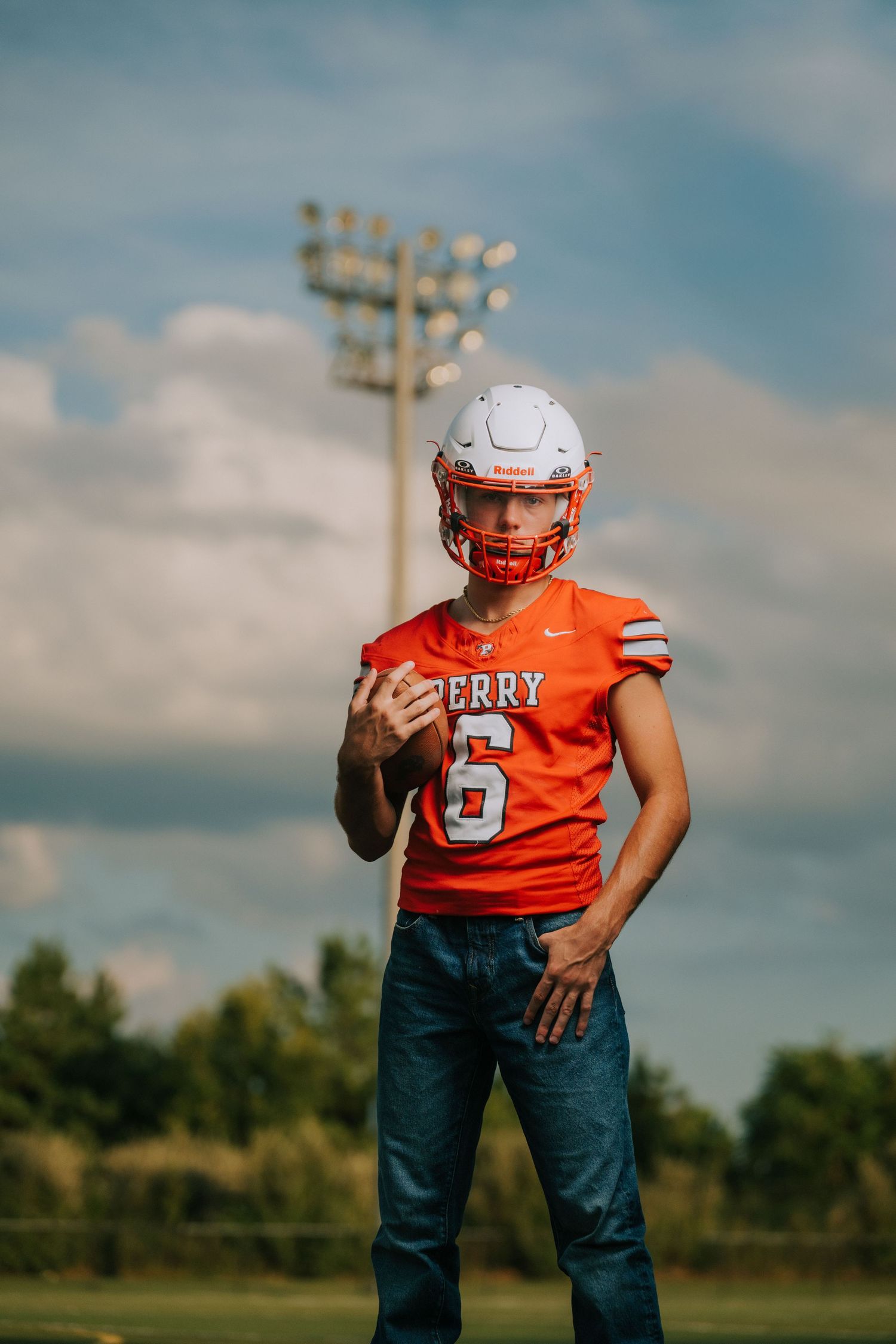 Football player in orange jersey and dark pants stands against cloudy sky with stadium lights in background.