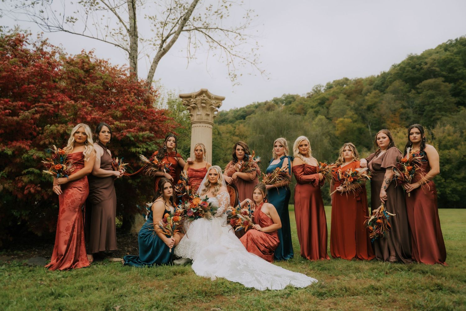 Bride in white gown poses with bridesmaids in rust, brown, and teal dresses holding fall bouquets outdoors.