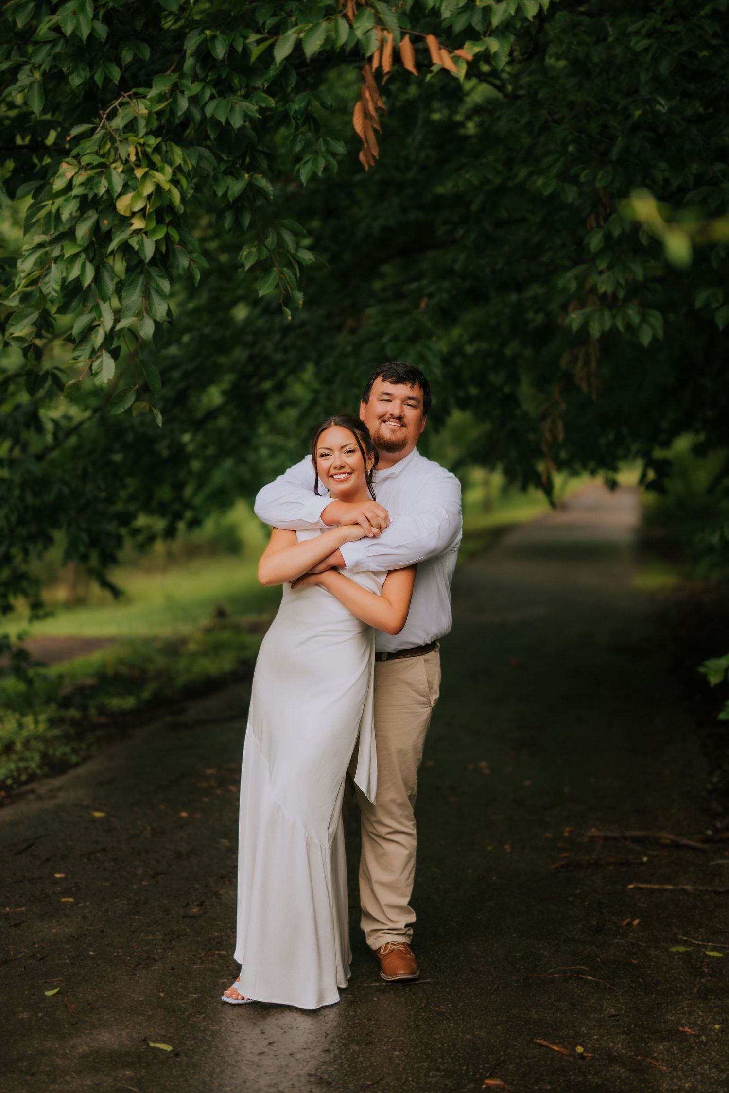 A couple embraces on a rain-soaked path surrounded by lush green trees, the woman in a white dress smiling joyfully.