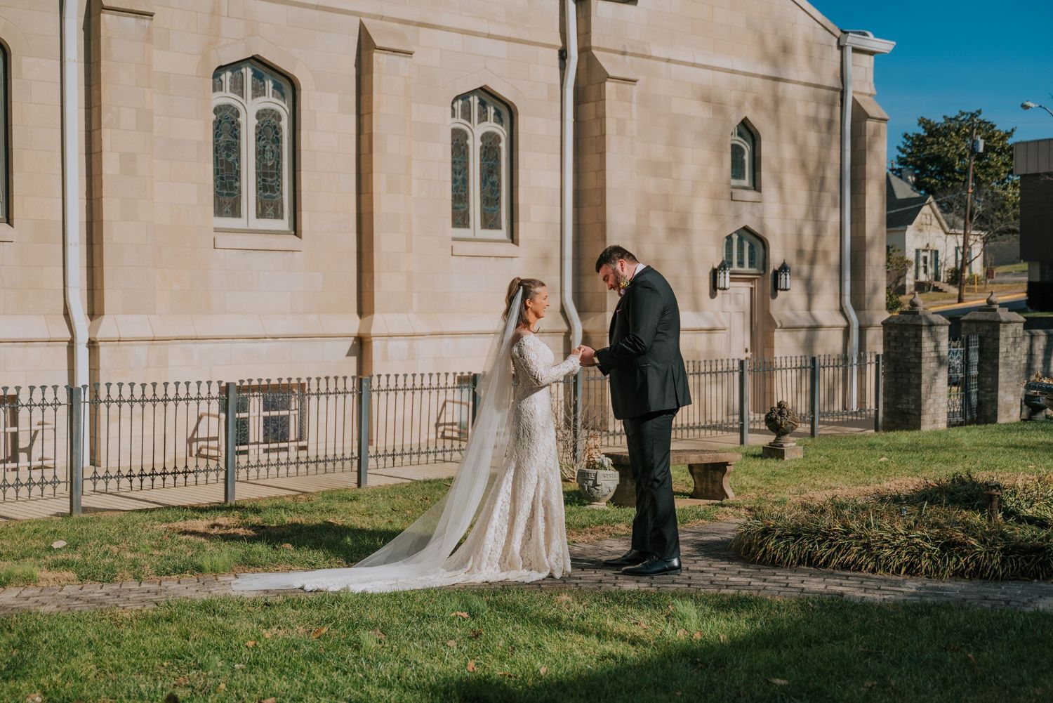 A wedding couple stands outside a stone church with arched windows on a sunny day.
