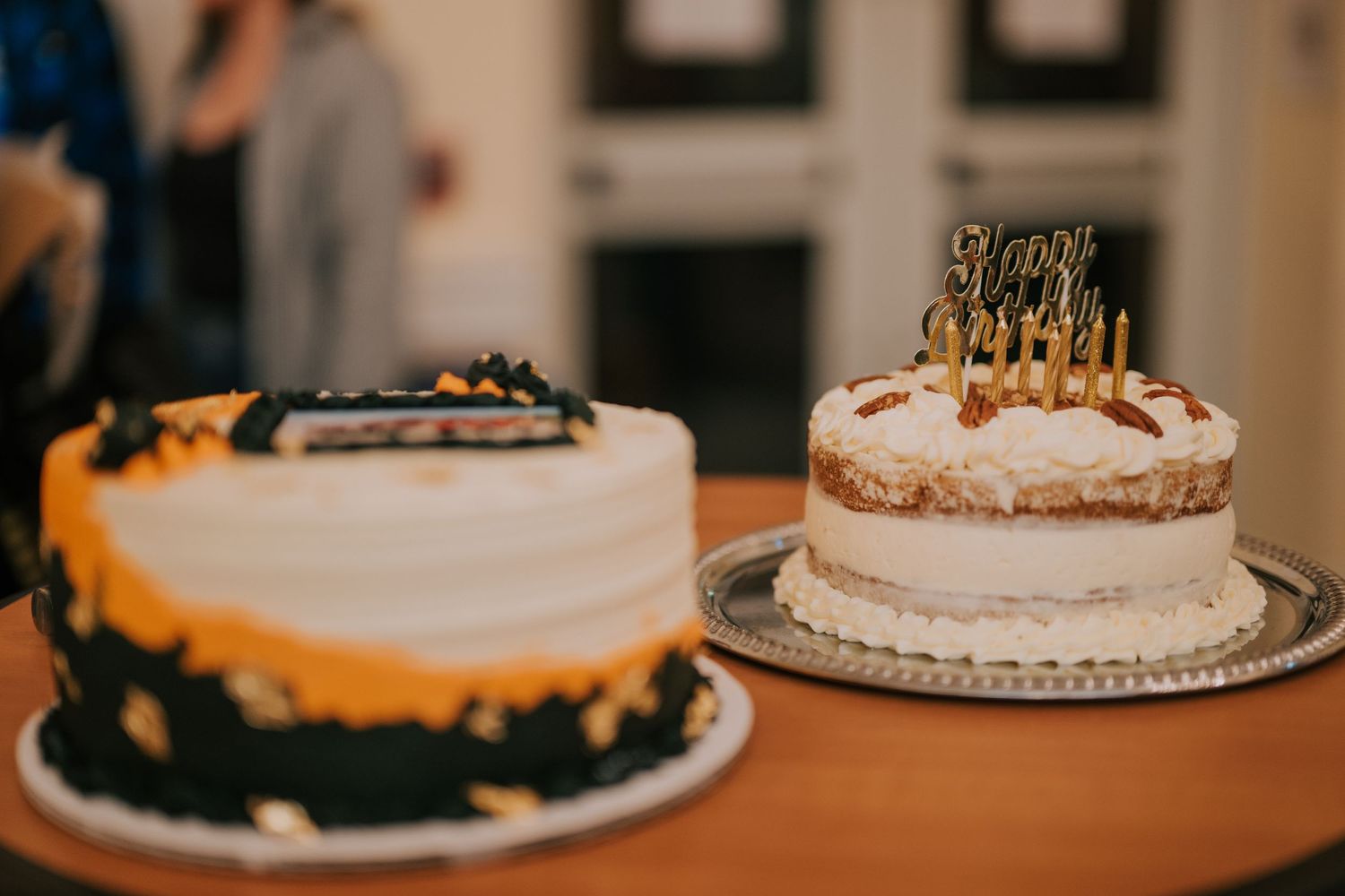Two elegant birthday cakes on wooden table with one decorated in black and white and another with golden birthday candles.